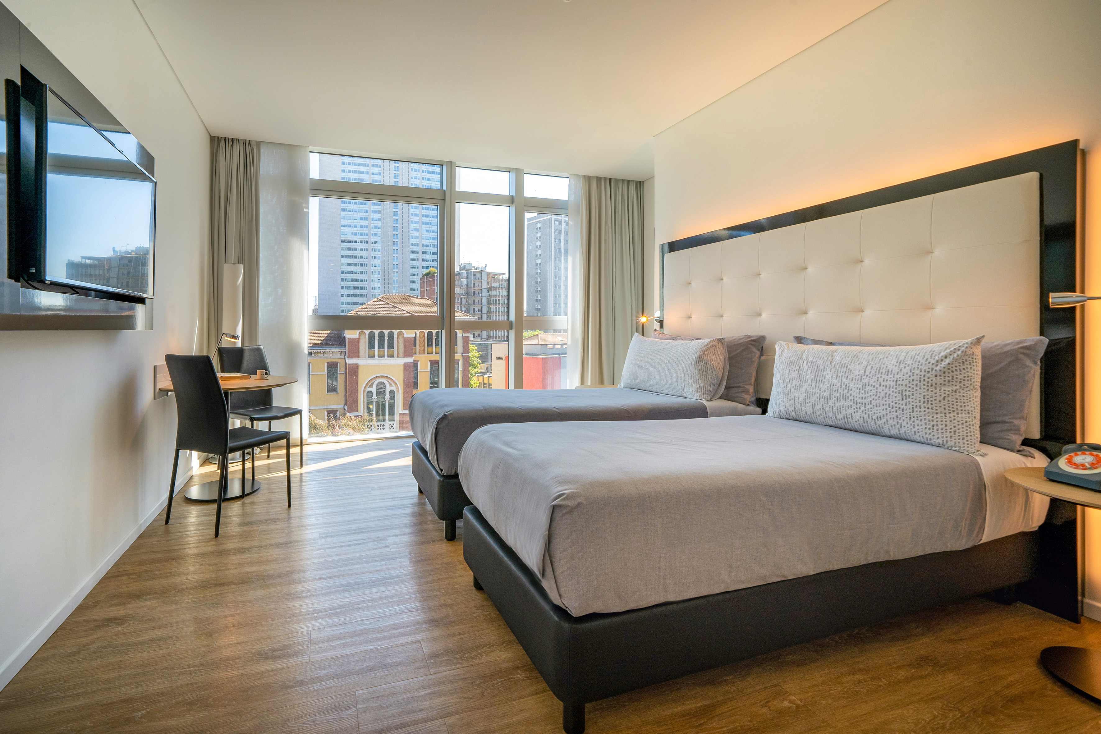 Modern hotel room with twin beds, city view, TV, and dining area.