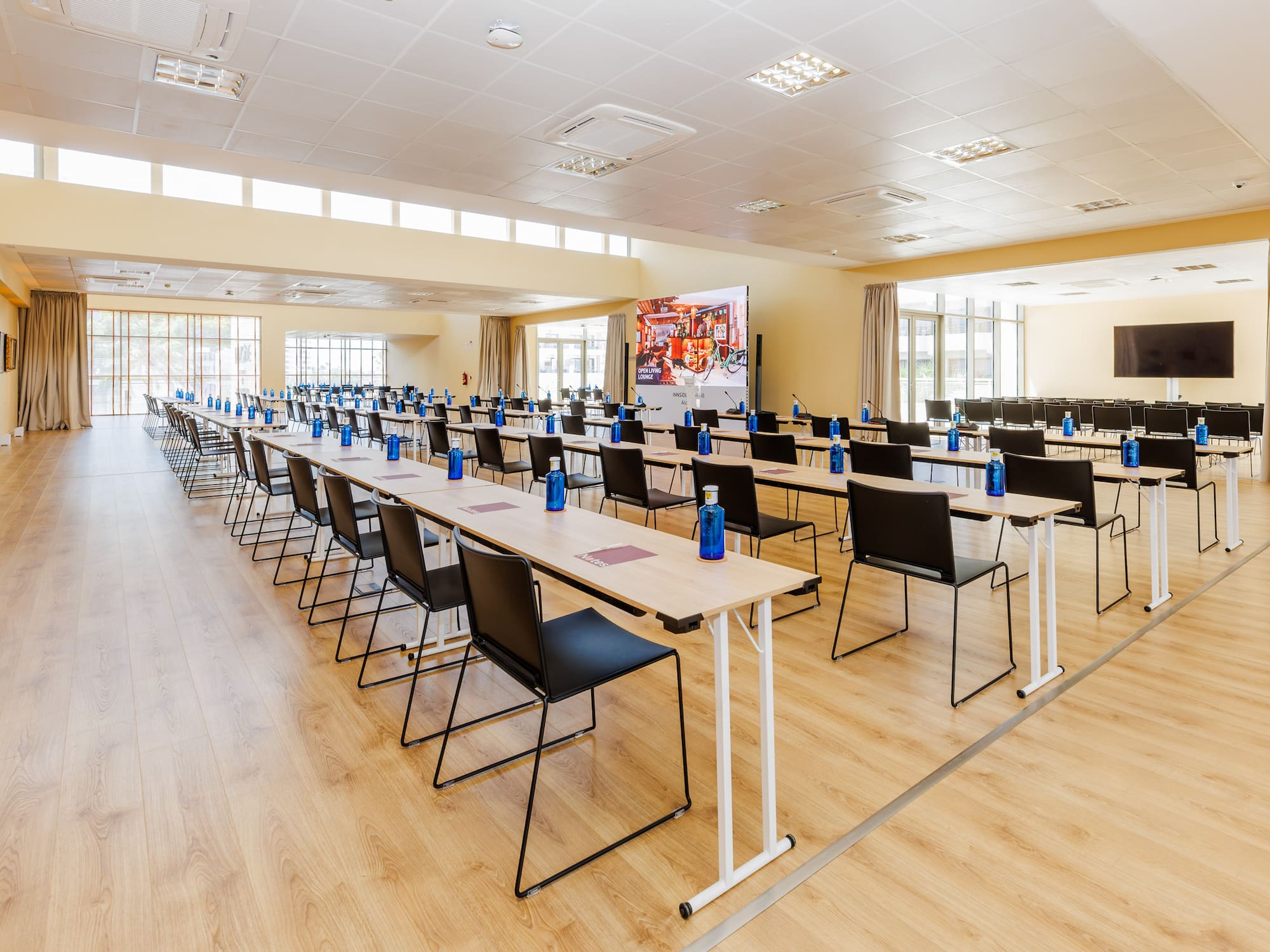 Modern conference room with tables, chairs, blue water bottles, large screen.