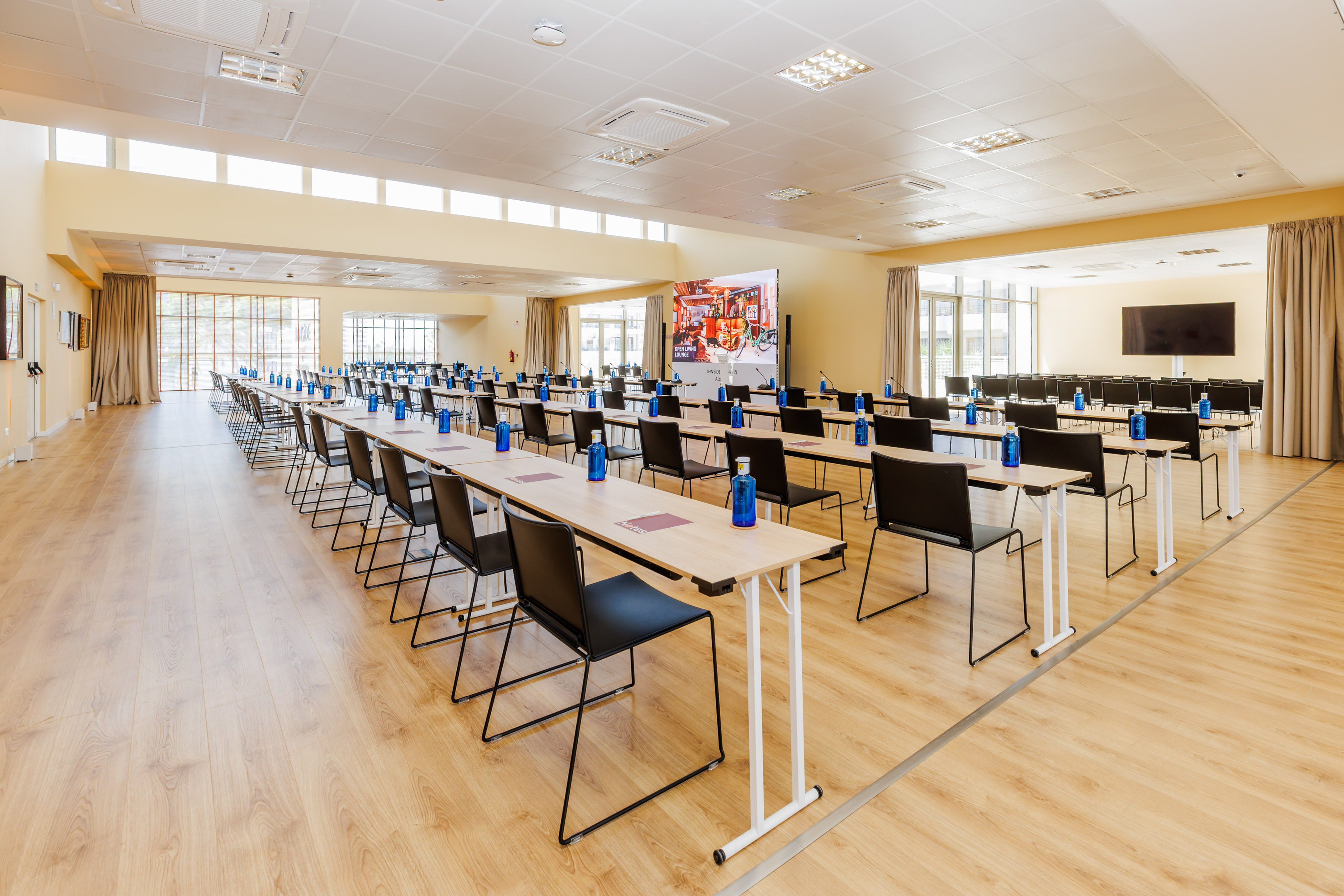 Modern conference room with tables, chairs, blue water bottles, large screen.
