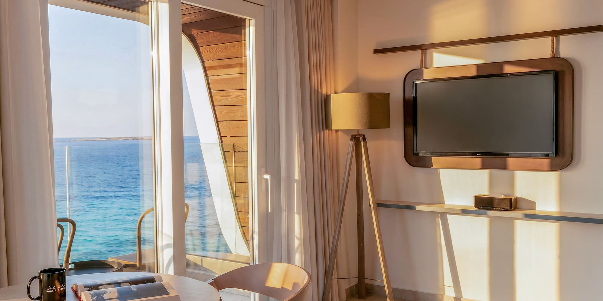 Waterfront room with sea view balcony, table, and TV in warm sunlight.