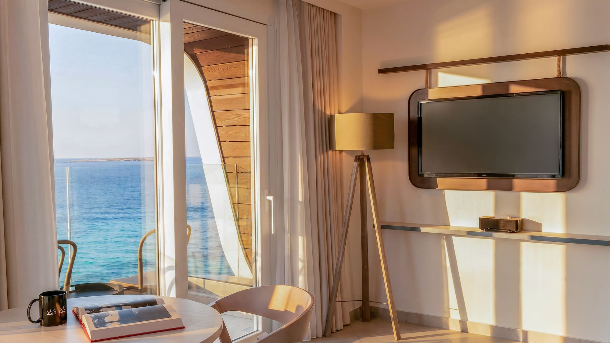 Waterfront room with sea view balcony, table, and TV in warm sunlight.