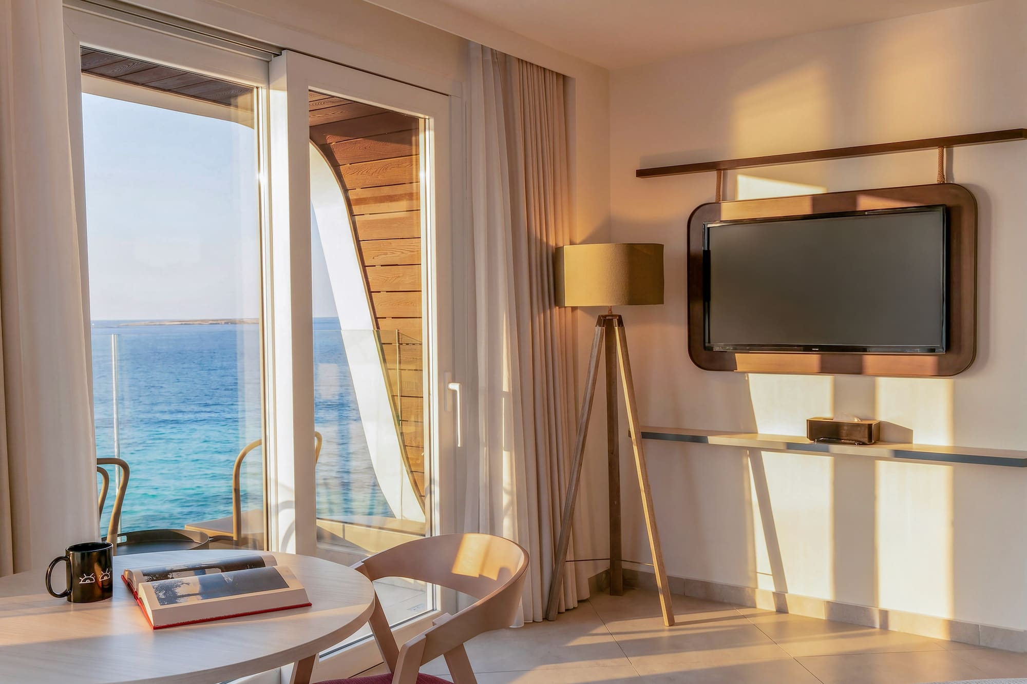 Waterfront room with sea view balcony, table, and TV in warm sunlight.