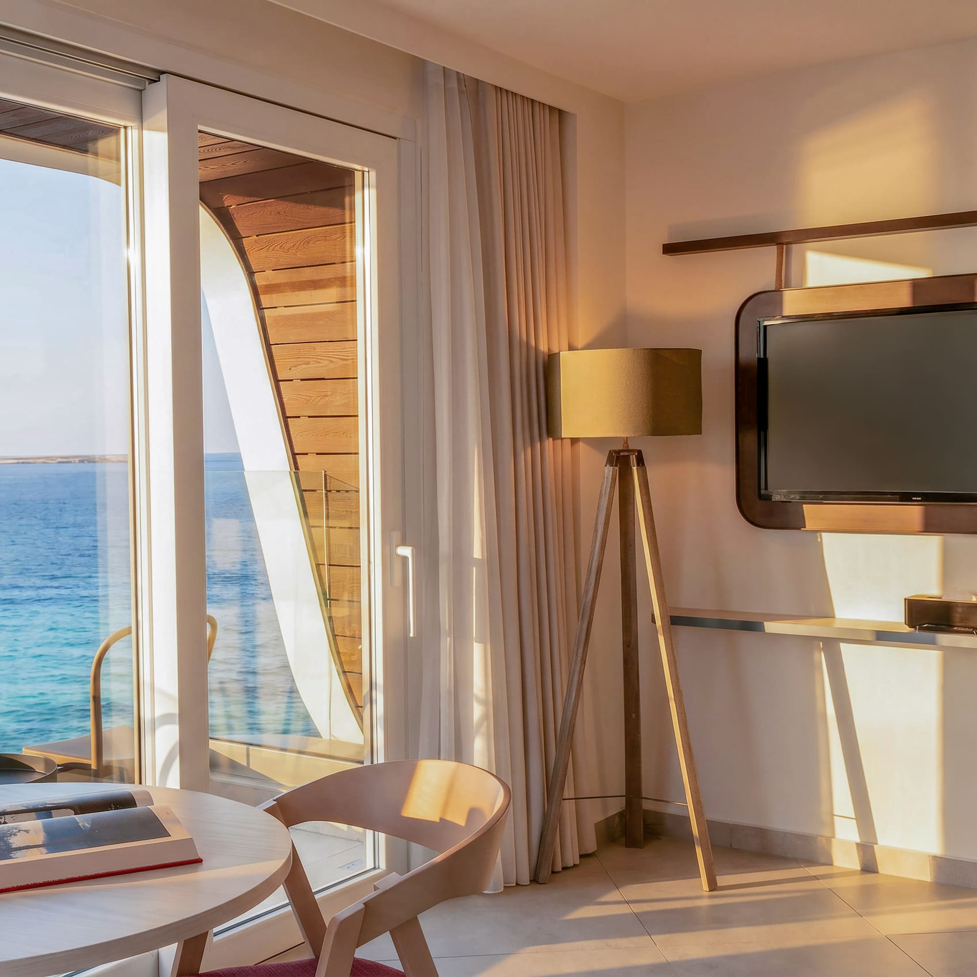Waterfront room with sea view balcony, table, and TV in warm sunlight.
