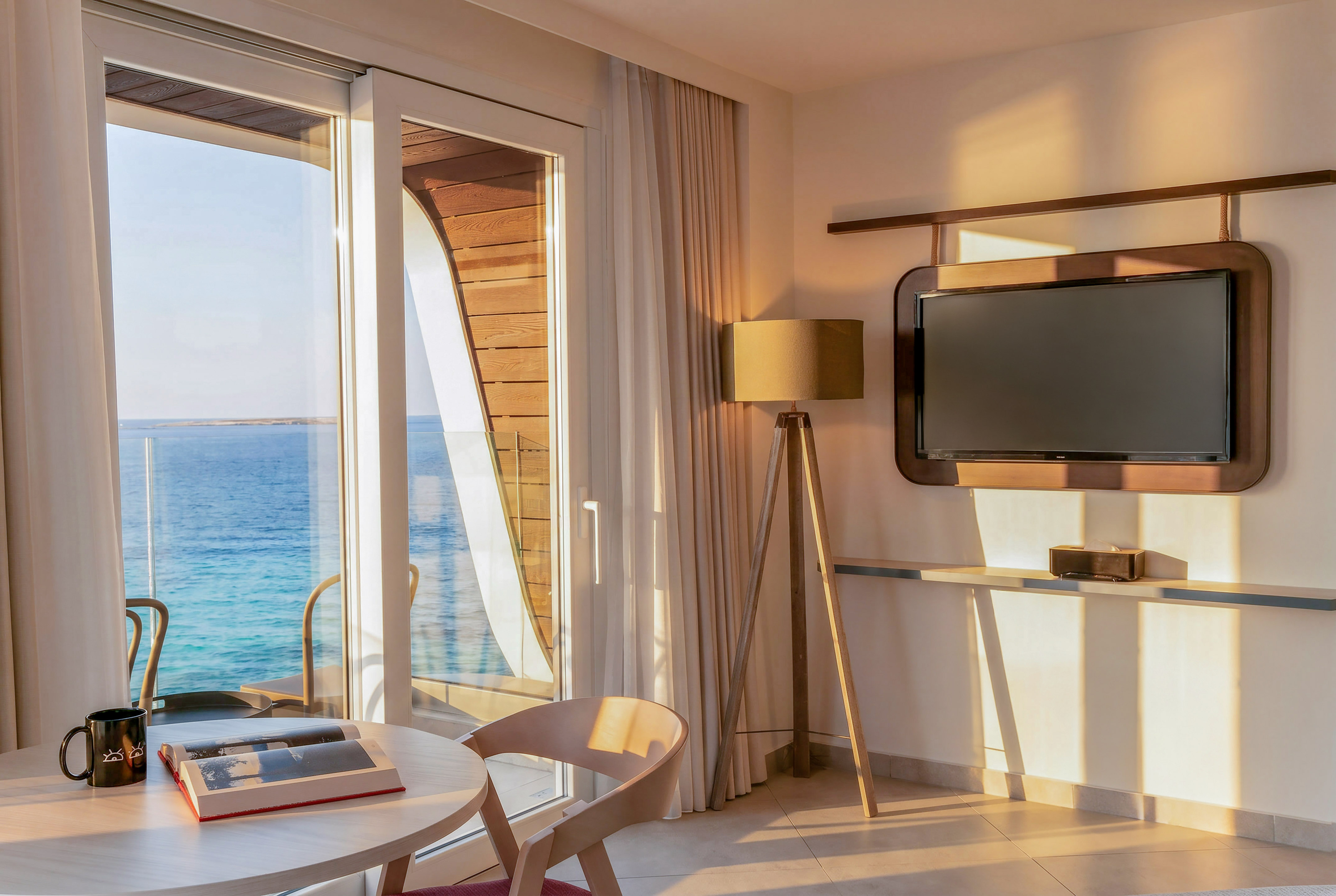Waterfront room with sea view balcony, table, and TV in warm sunlight.