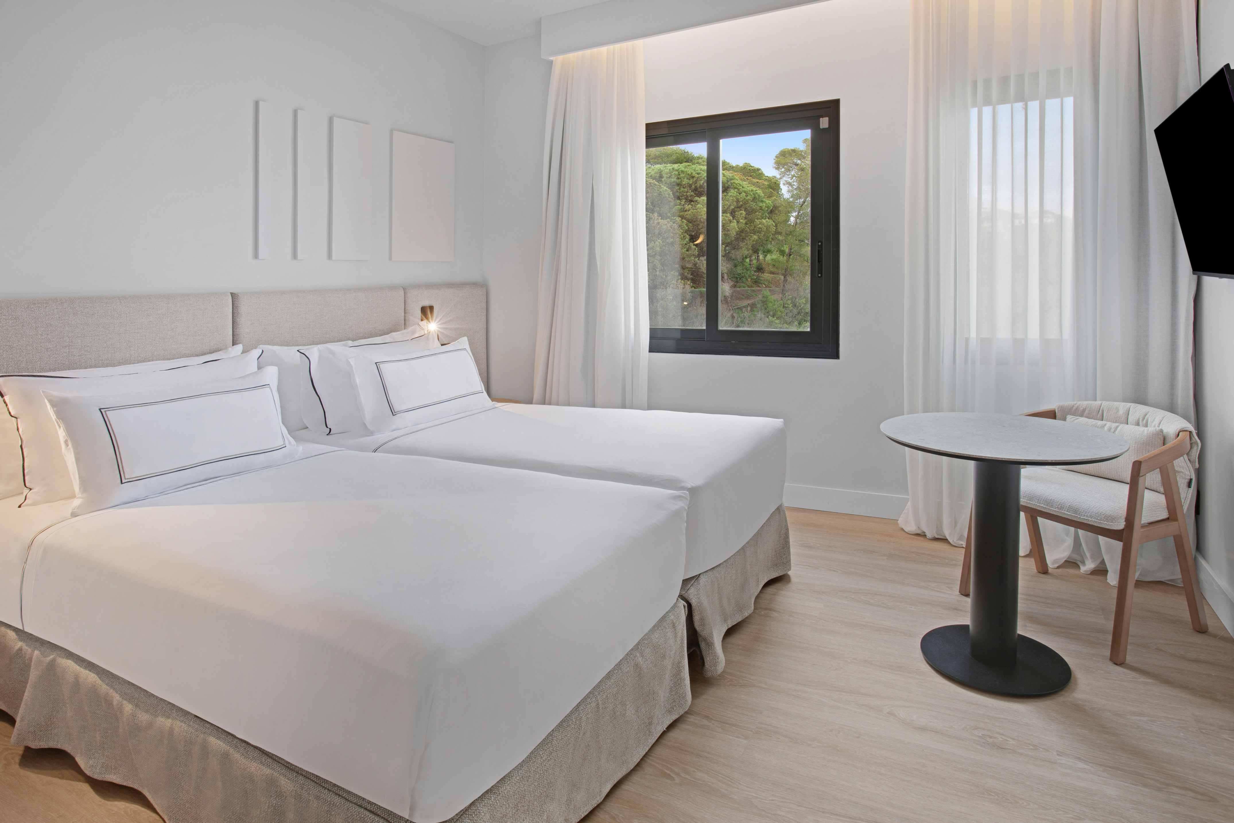 Modern hotel twin room with two white beds, window with green view, light wood floor.