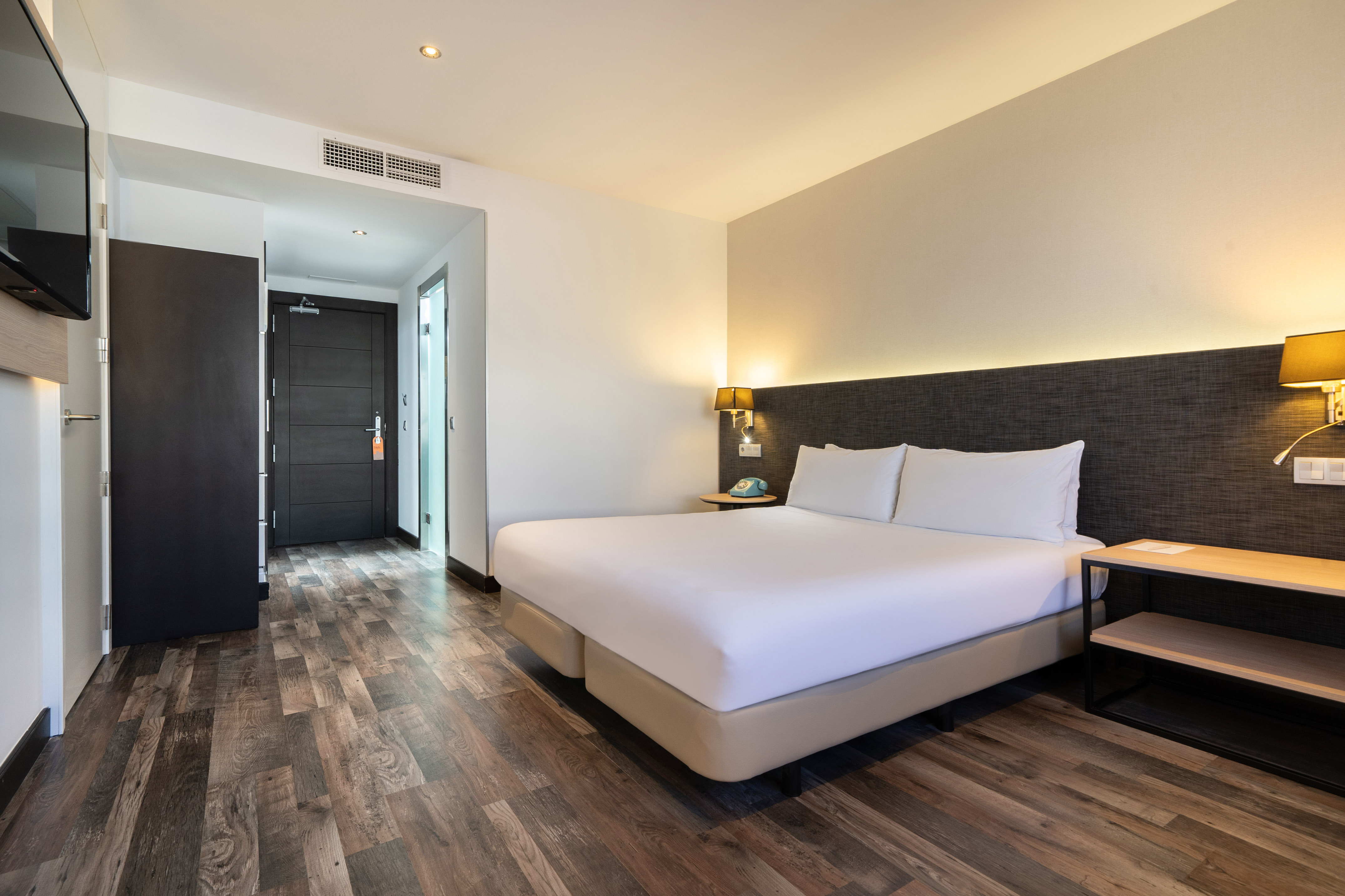 Modern hotel room with king bed, dark wood floor, and minimalist decor.