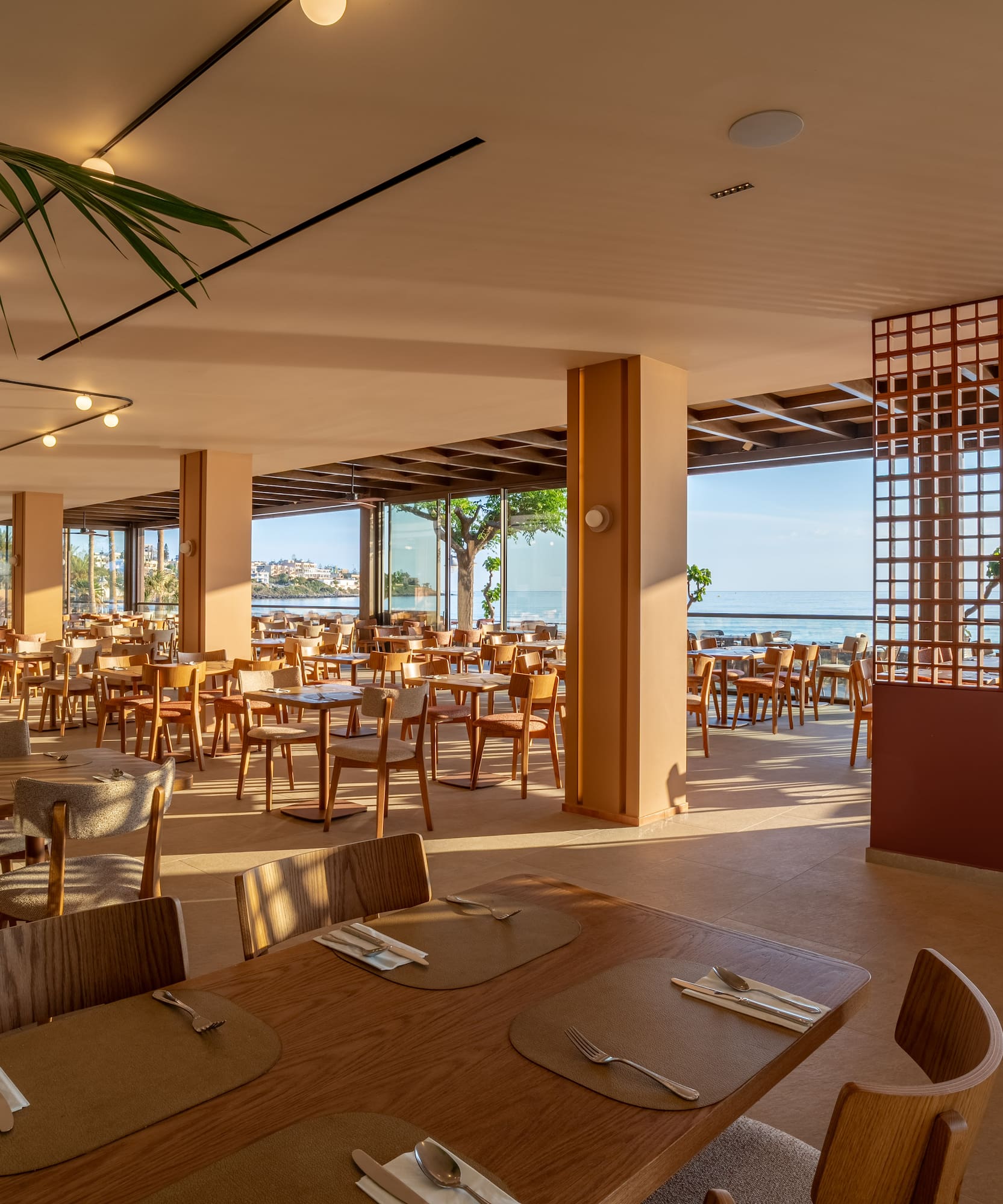 Sunny modern hotel restaurant with ocean view, set dining tables, and lush plants.