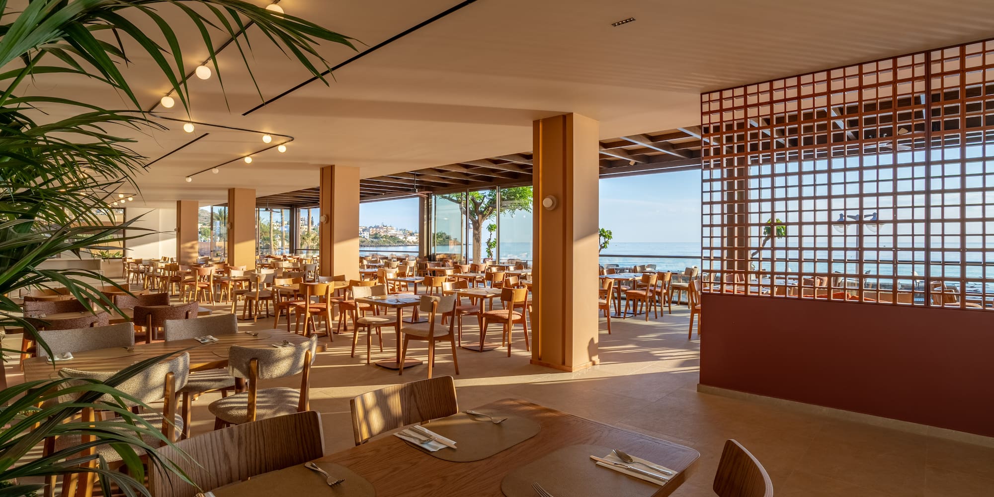 Spacious restaurant with sea view, wooden dining tables, and tropical plants.