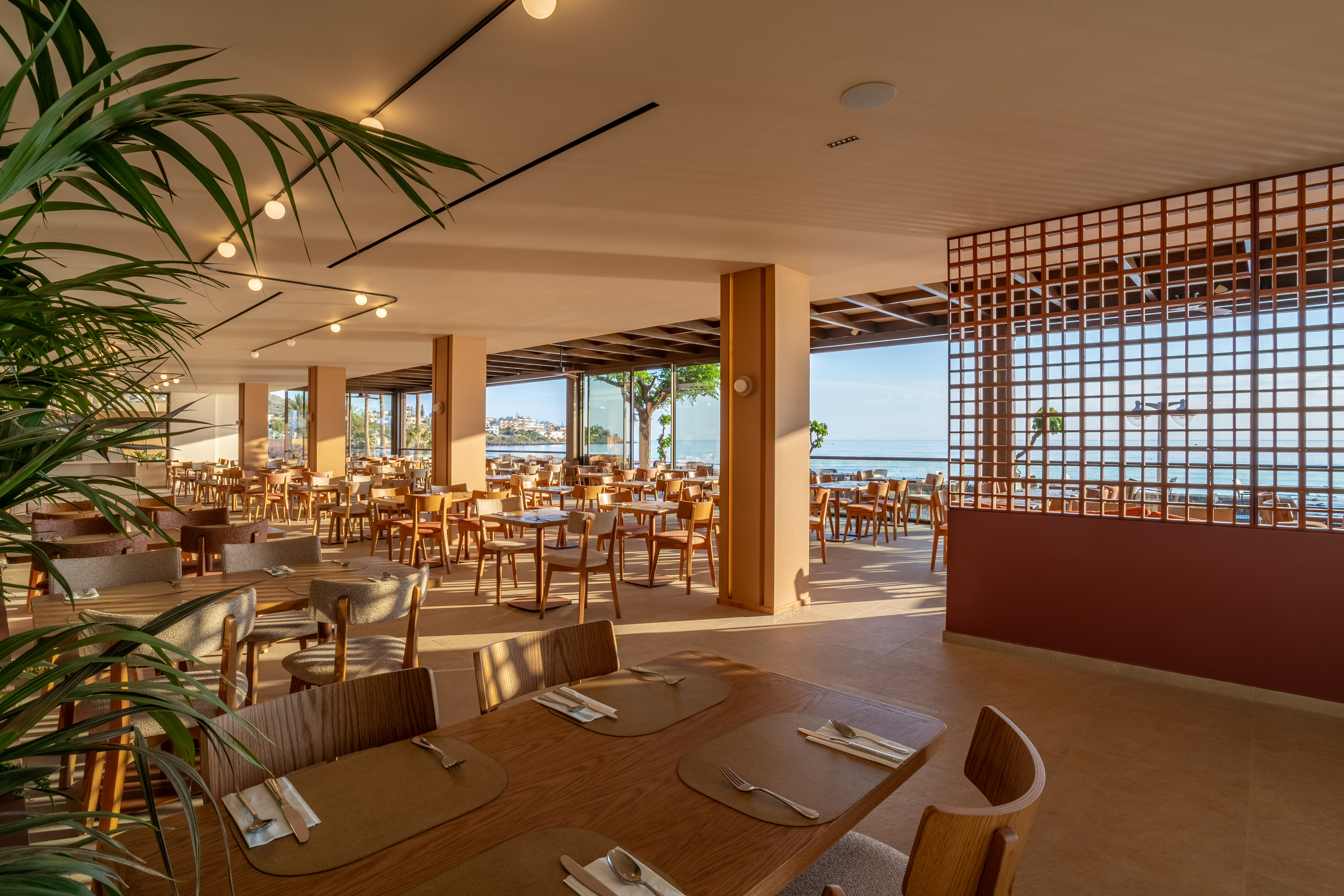 Sunny modern hotel restaurant with ocean view, set dining tables, and lush plants.