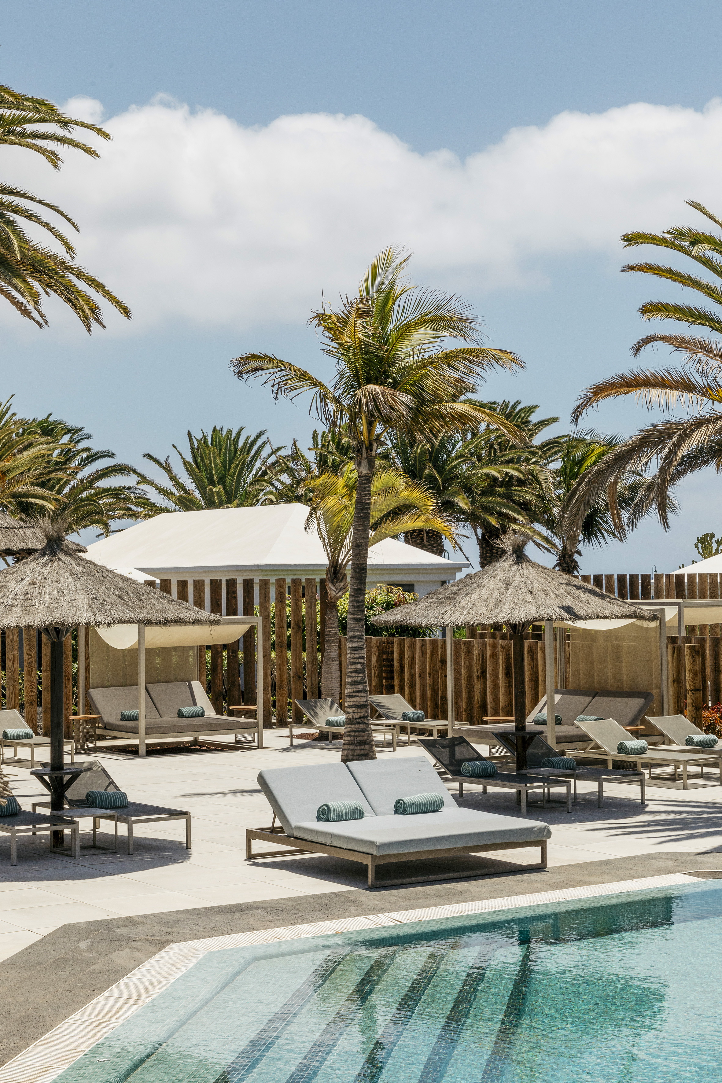 Resort swimming pool with sunbeds, cabanas, and tropical palm trees.