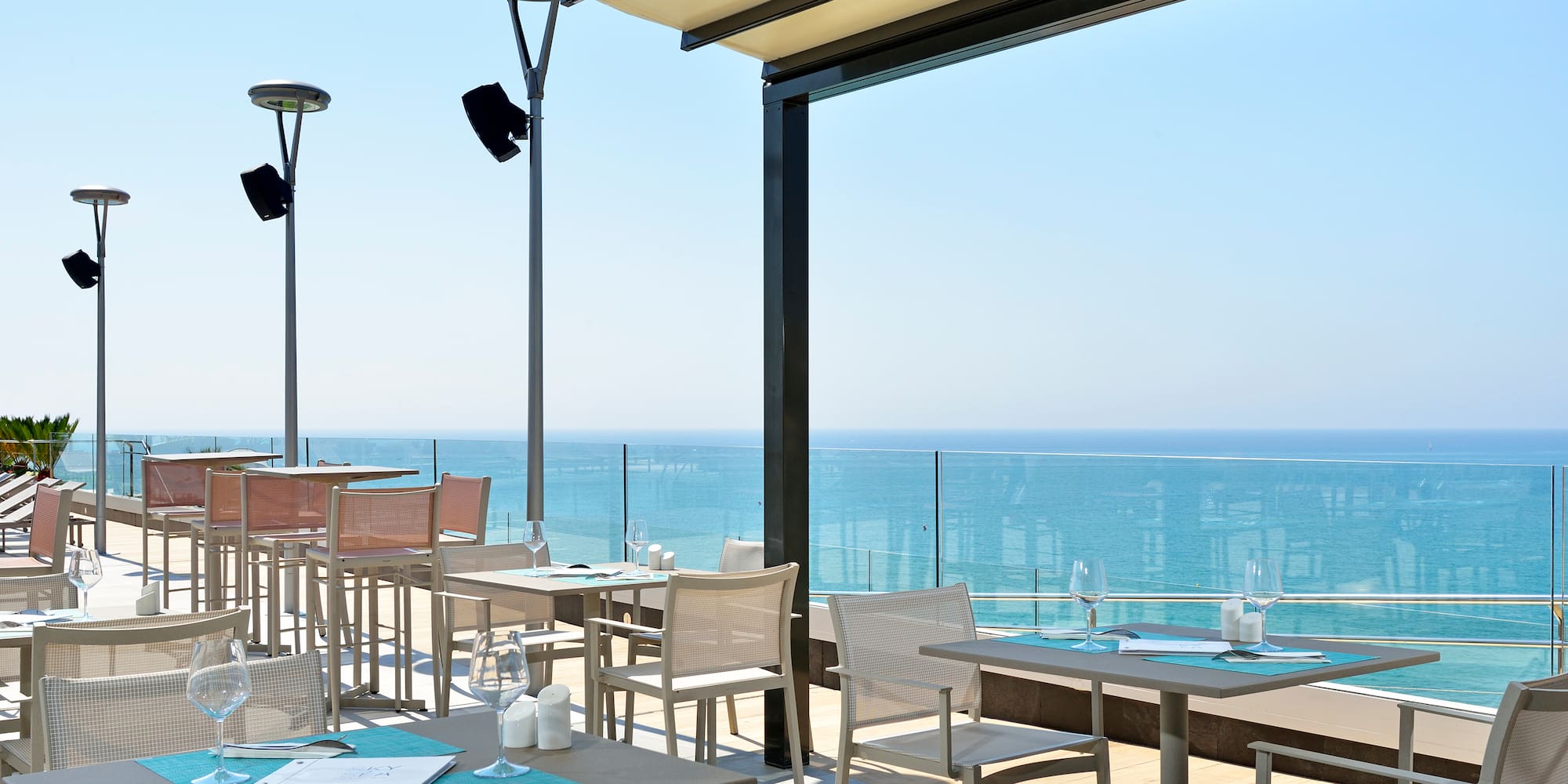 Sunny outdoor restaurant terrace with ocean view and dining tables.