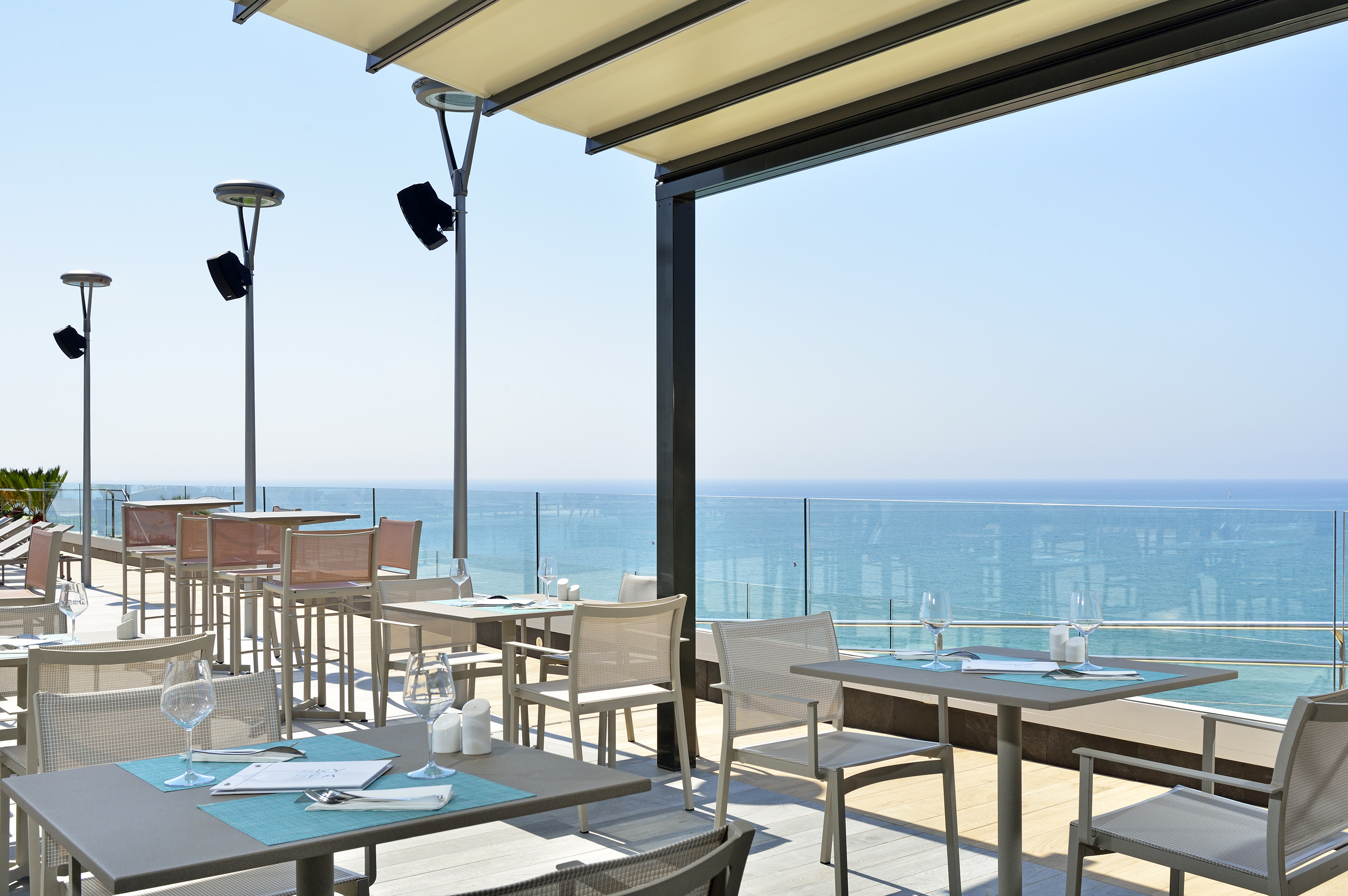 Sunny outdoor restaurant terrace with ocean view and dining tables.