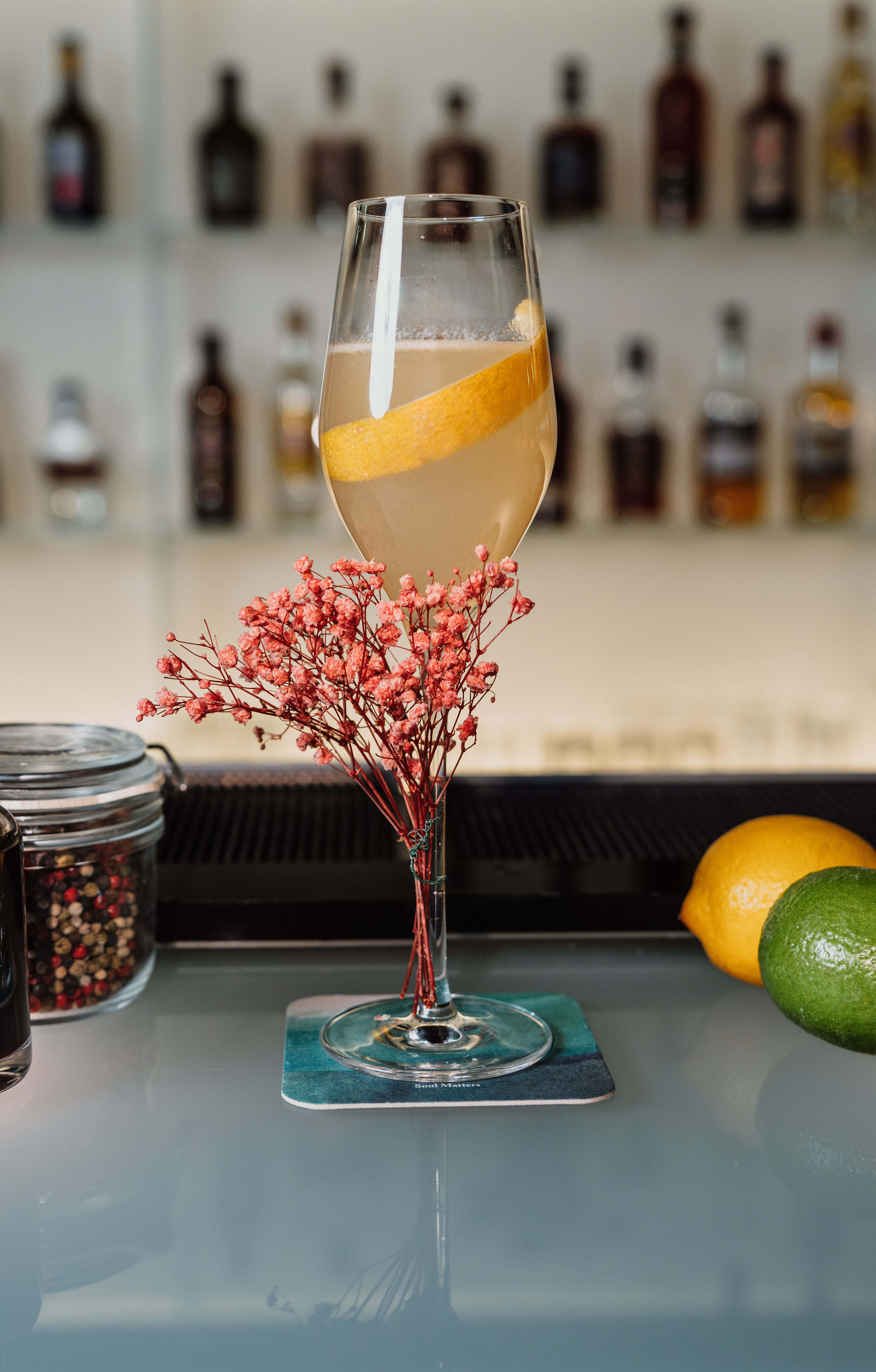 Cocktail with lemon peel, pink flowers, lemon, lime, and bar background.