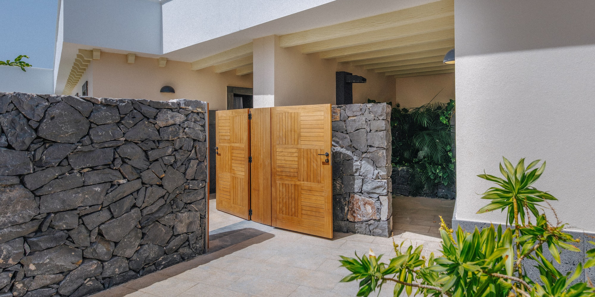 Stone wall and open wooden gate leading to a bright modern villa entrance.