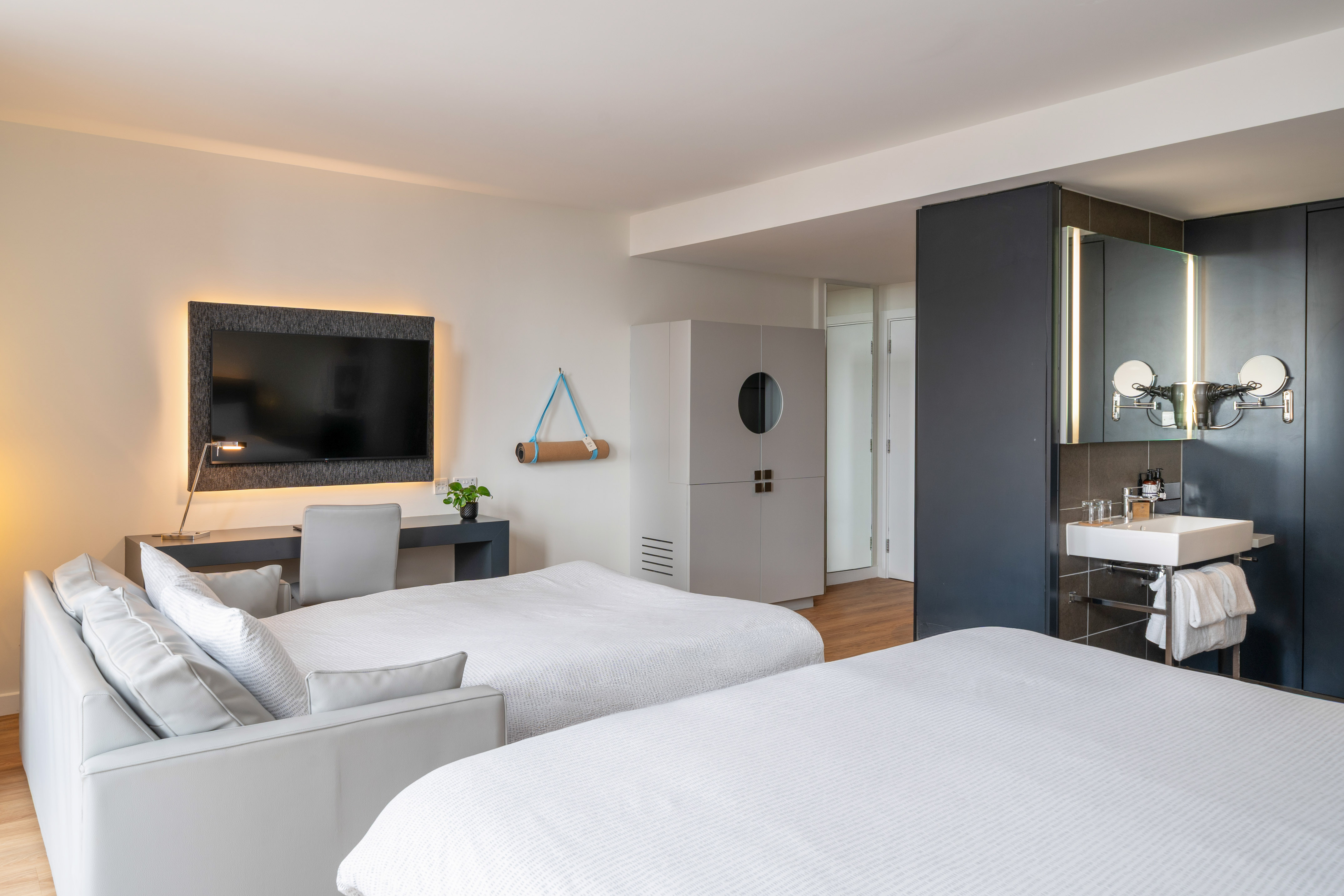 Modern hotel room with two beds, sofa, desk, backlit TV, yoga mat, and vanity.