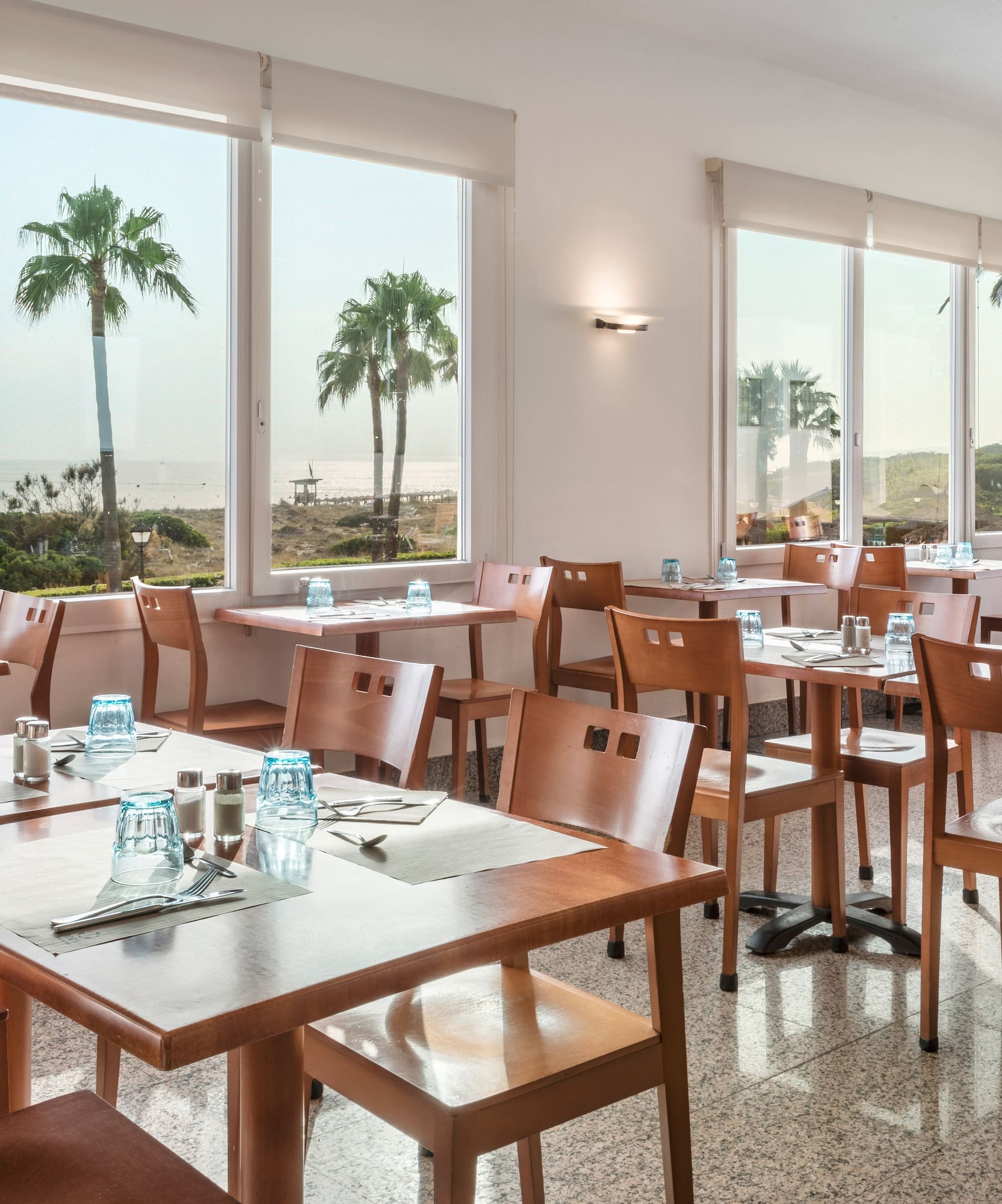 Restaurant dining room overlooking the ocean and palm trees, with set tables.