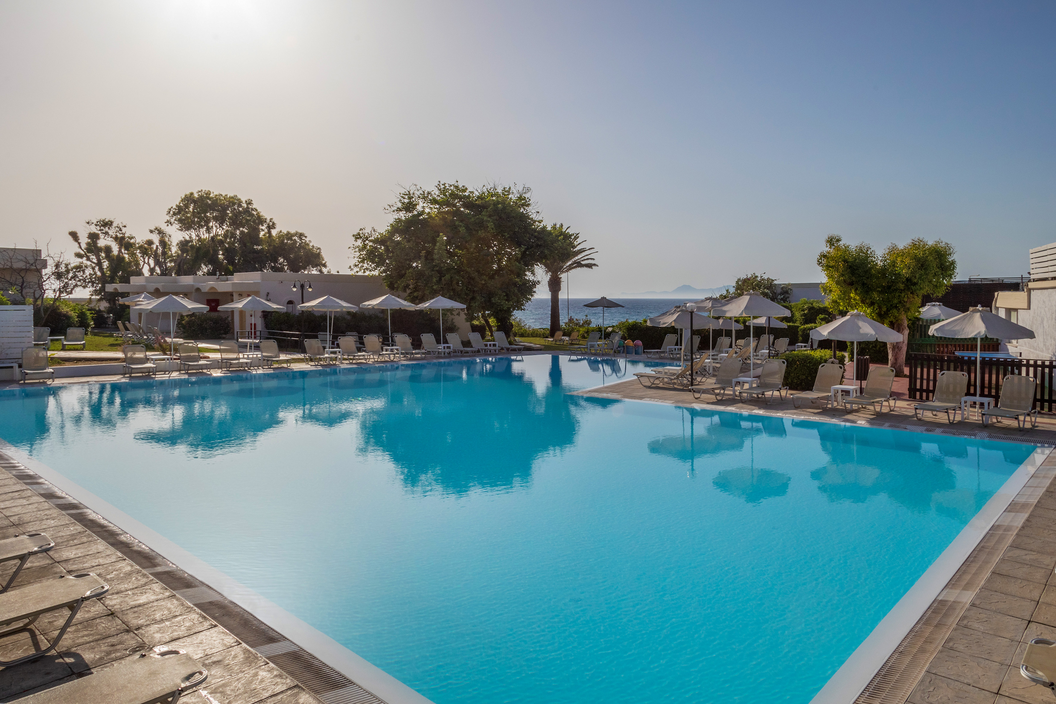 Sunny blue resort swimming pool with loungers, umbrellas, and distant sea view.