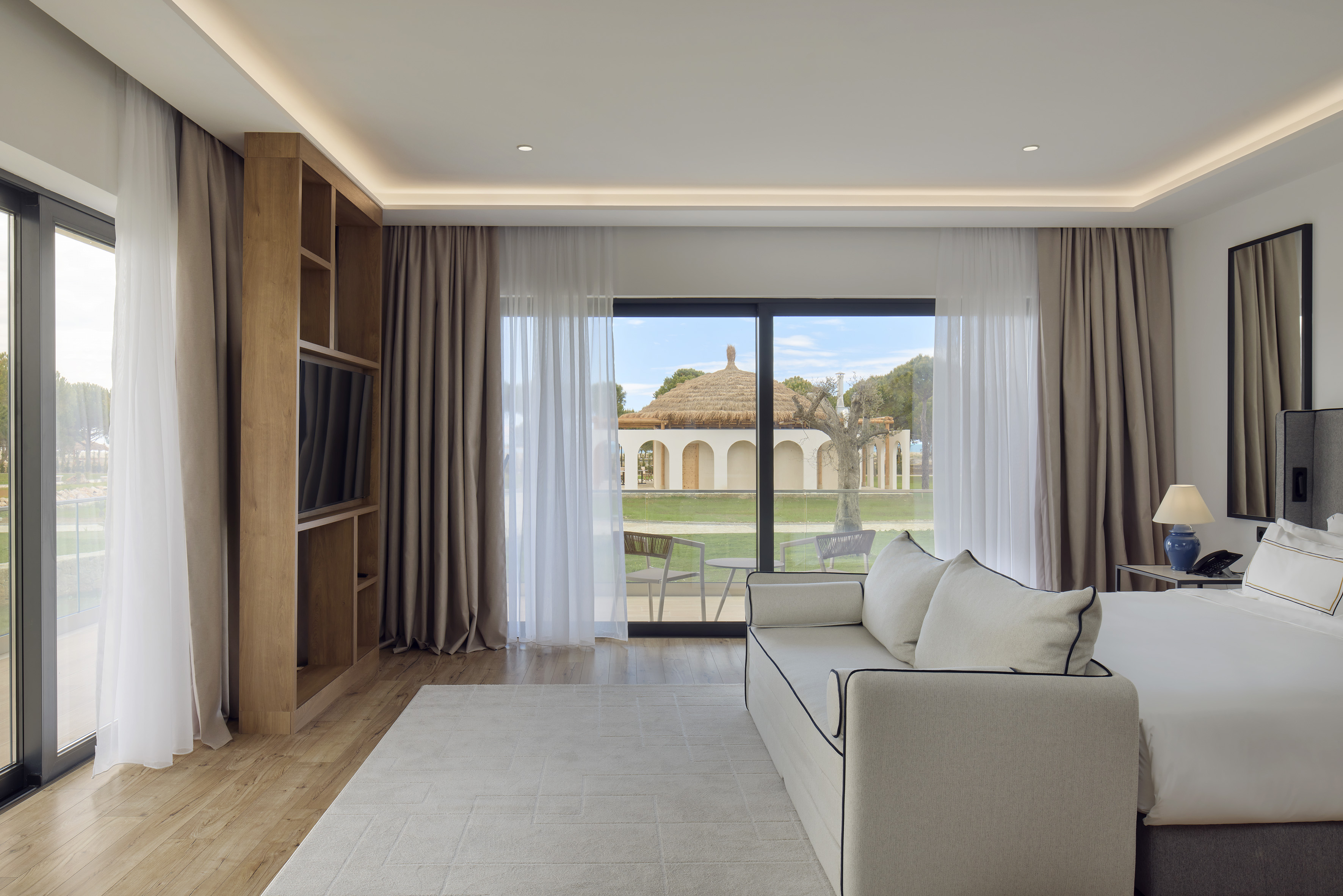 Modern hotel suite with bed, sofa, and garden view of thatched roof building.