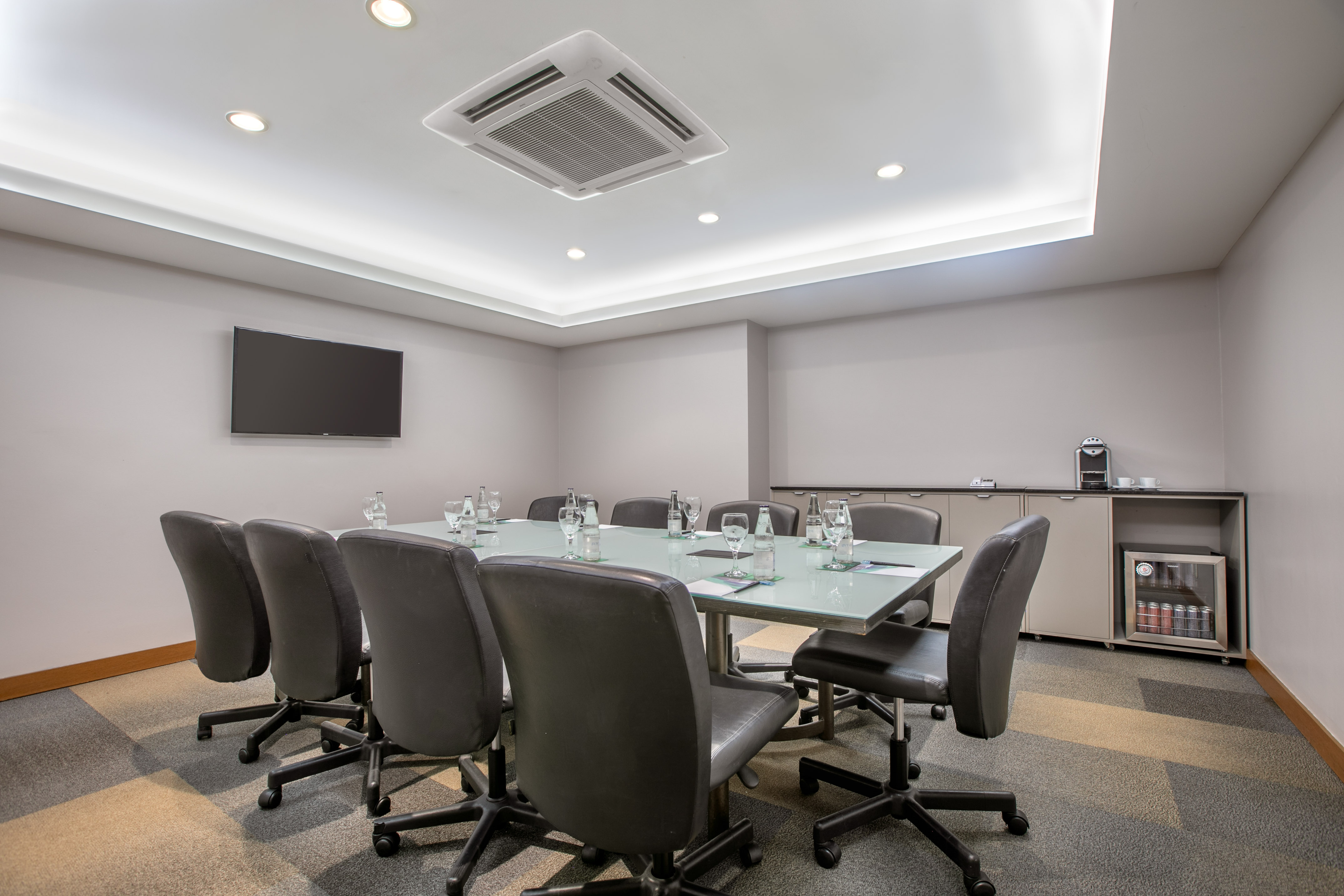 Modern conference room with glass table, office chairs, wall TV, and coffee station.