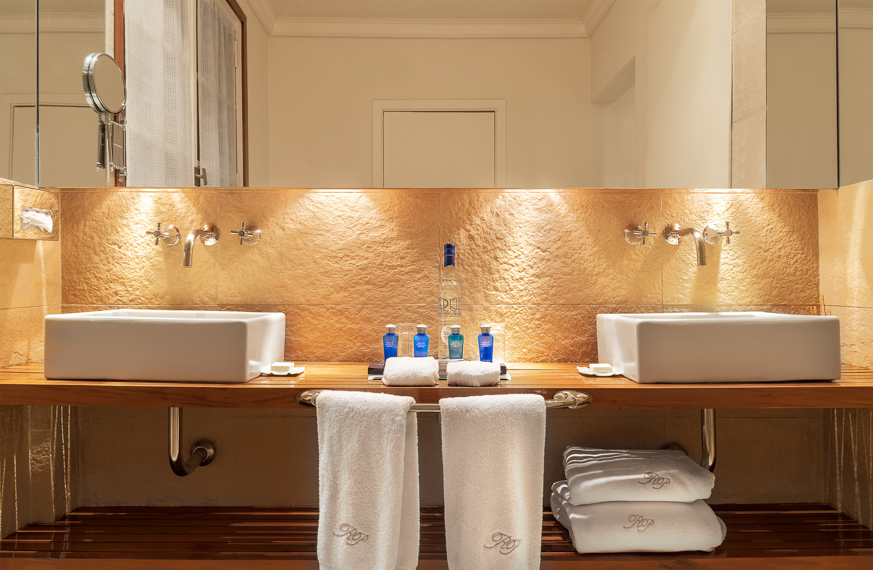 Double vanity bathroom: white sinks, wood counter, gold tiled wall, amenities.