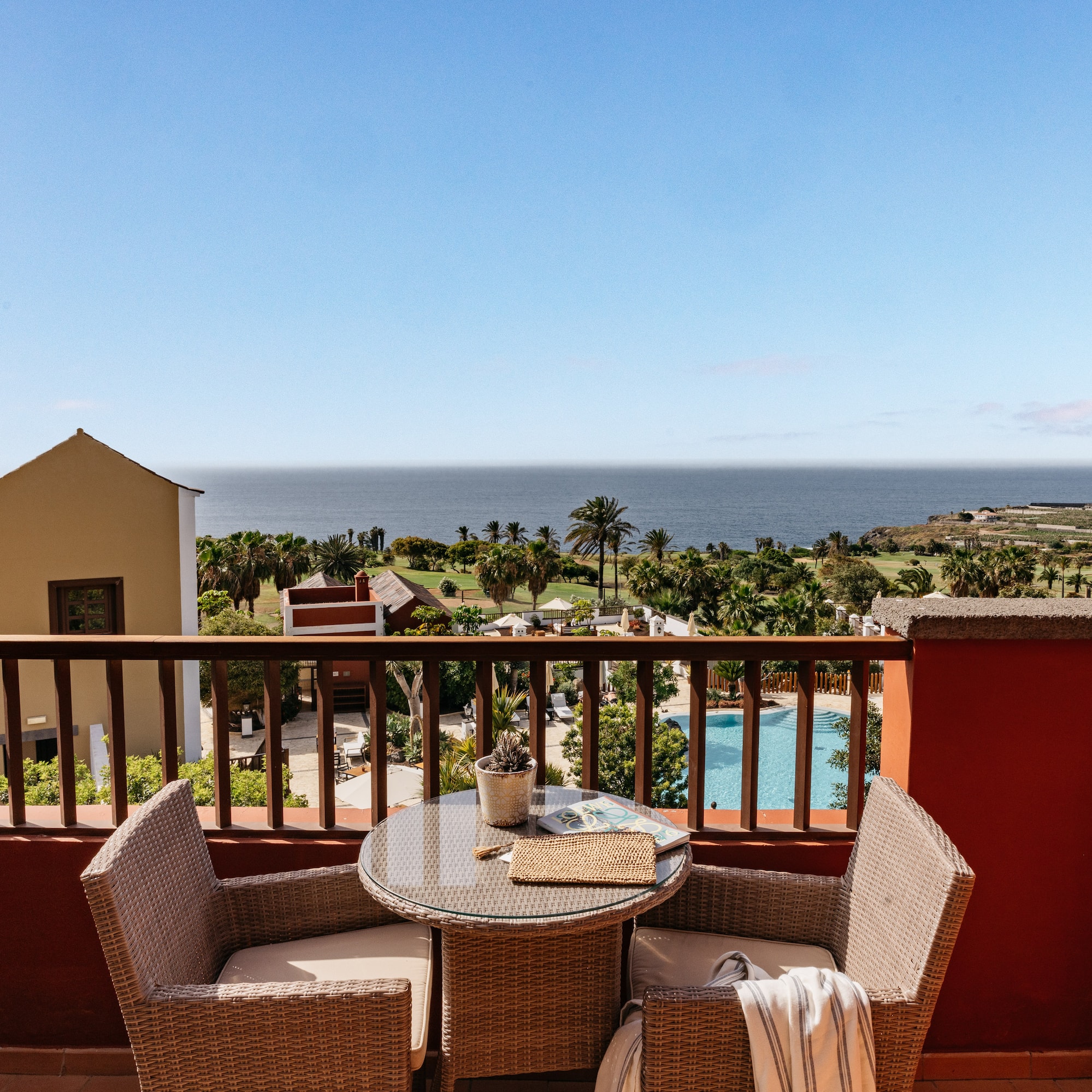 Oceanfront balcony with wicker furniture, palm trees, and pool view.