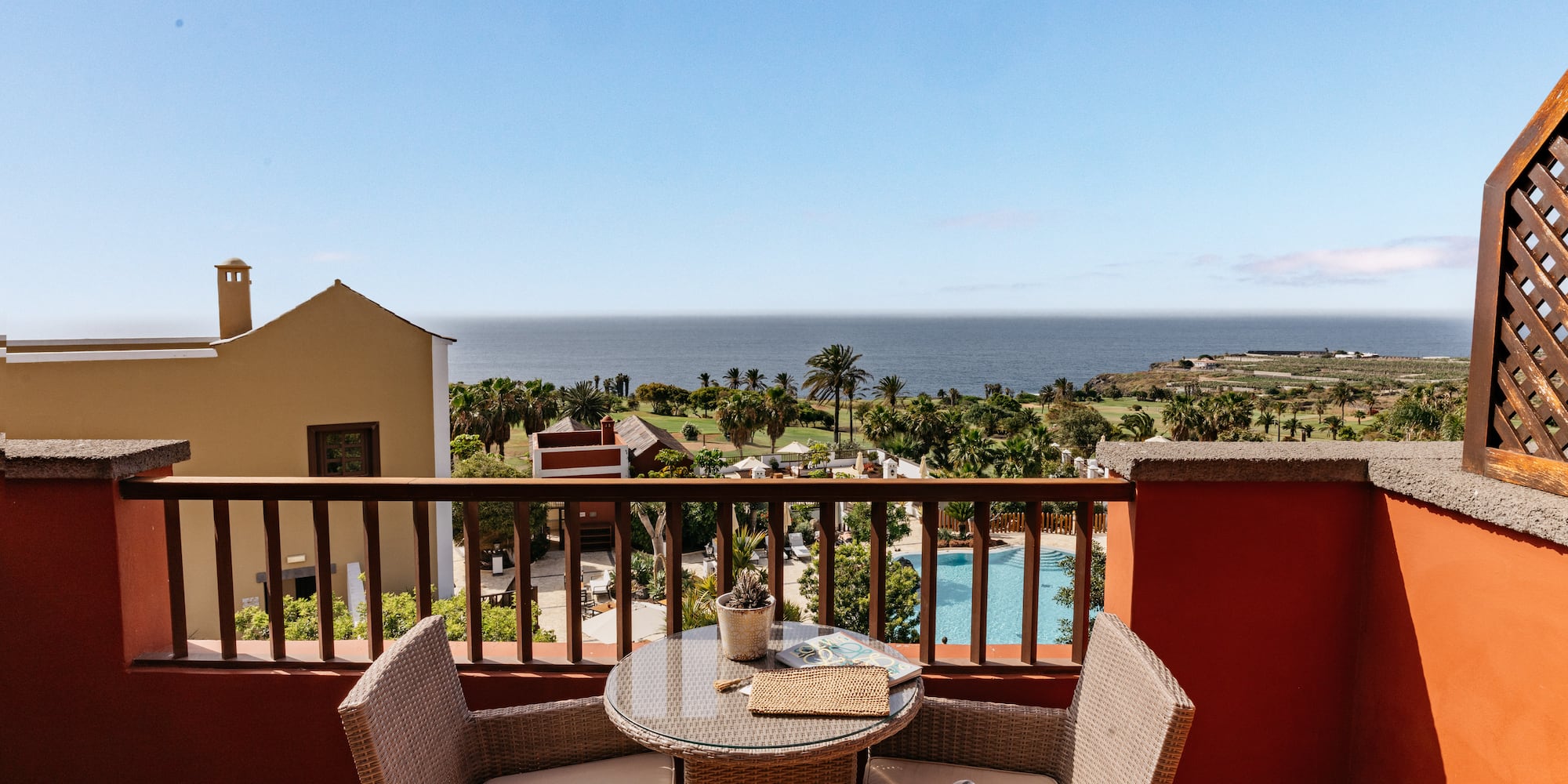 Balcony ocean view: wicker table, chairs, palm trees, and resort pool.