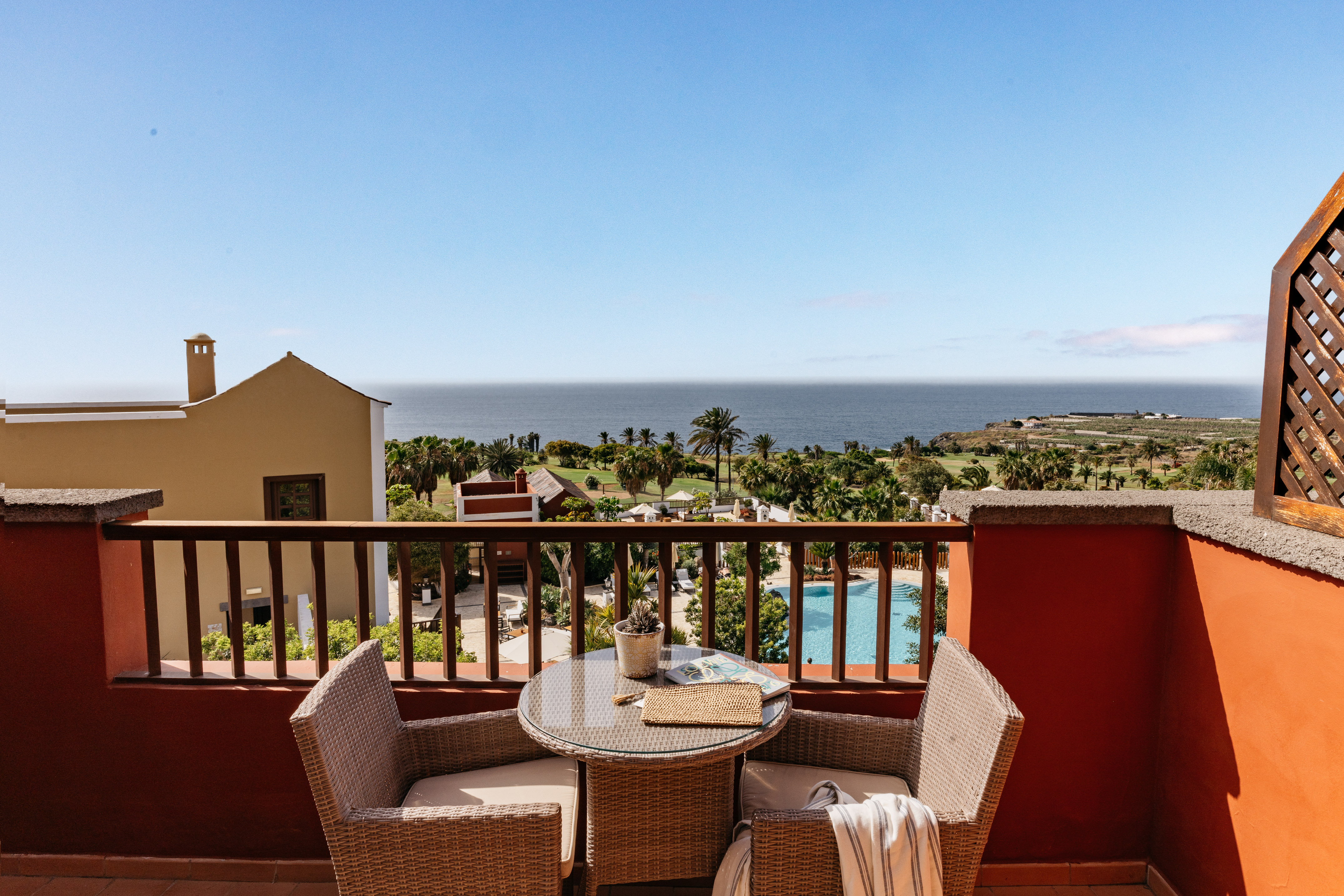 Oceanfront balcony with wicker furniture, palm trees, and pool view.