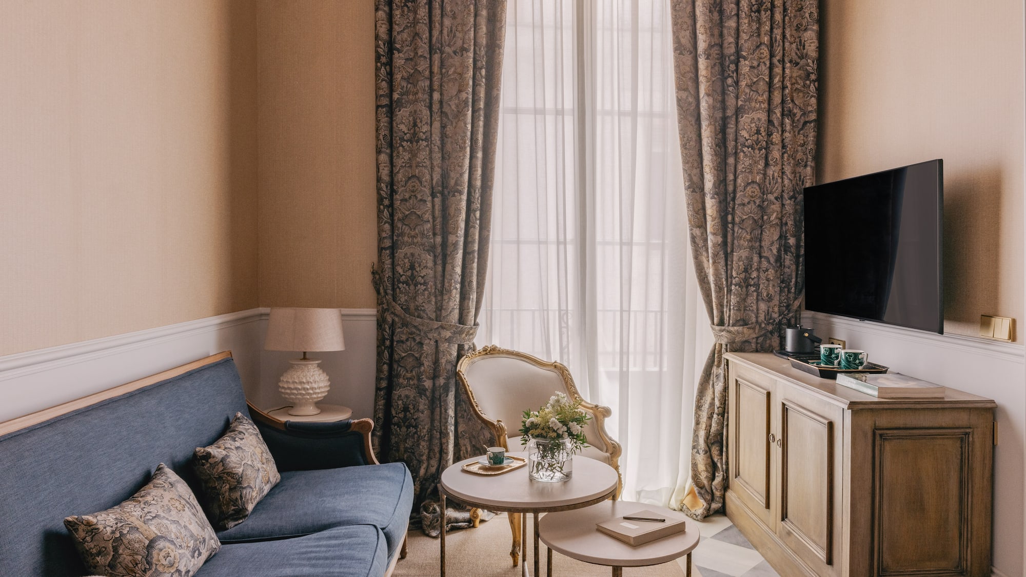 Acogedor salón de hotel con sofá azul, TV y cortinas elegantes.