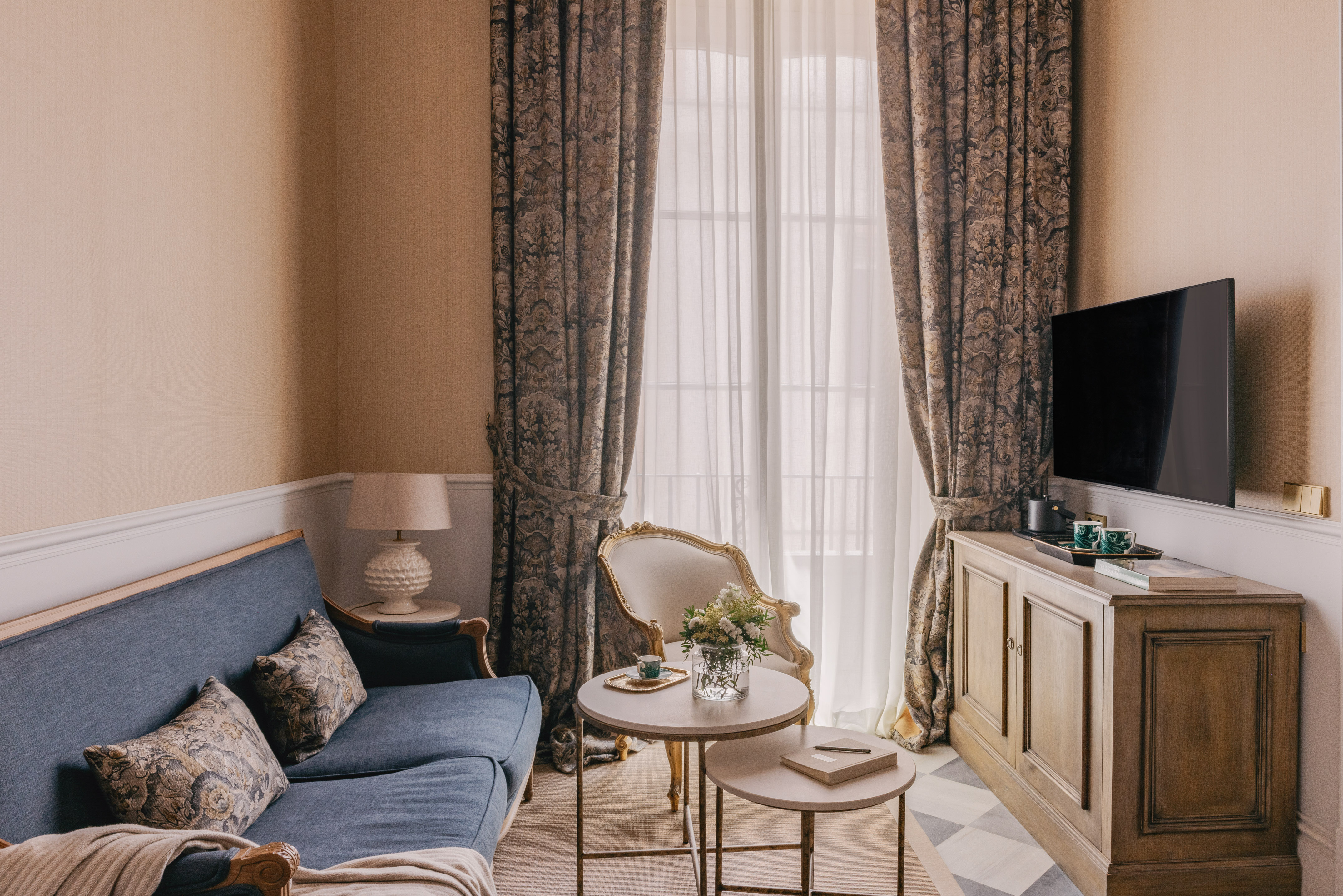 Acogedor salón de hotel con sofá azul, TV y cortinas elegantes.