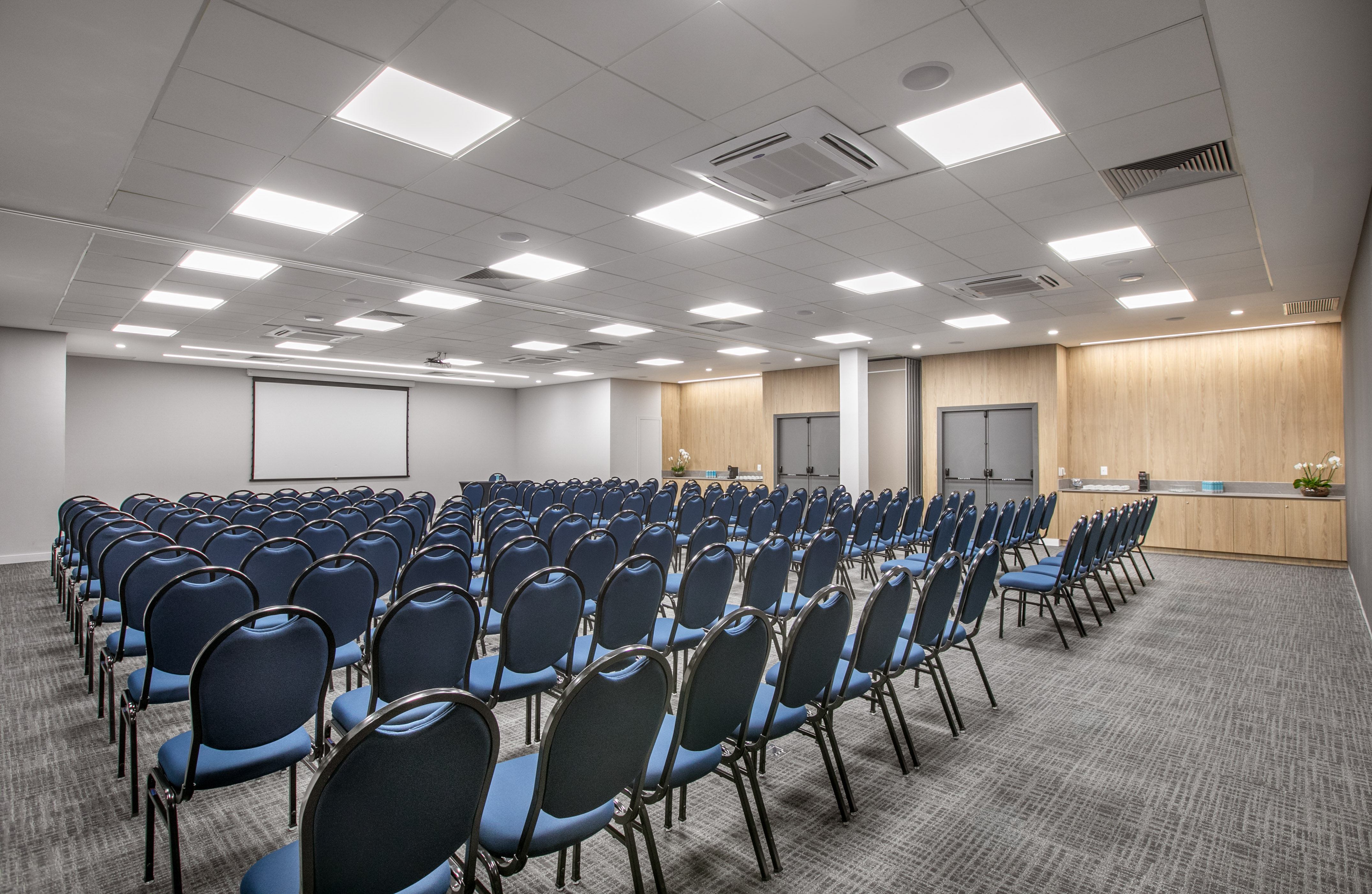 Empty conference room with rows of blue chairs, projection screen, and wood details.