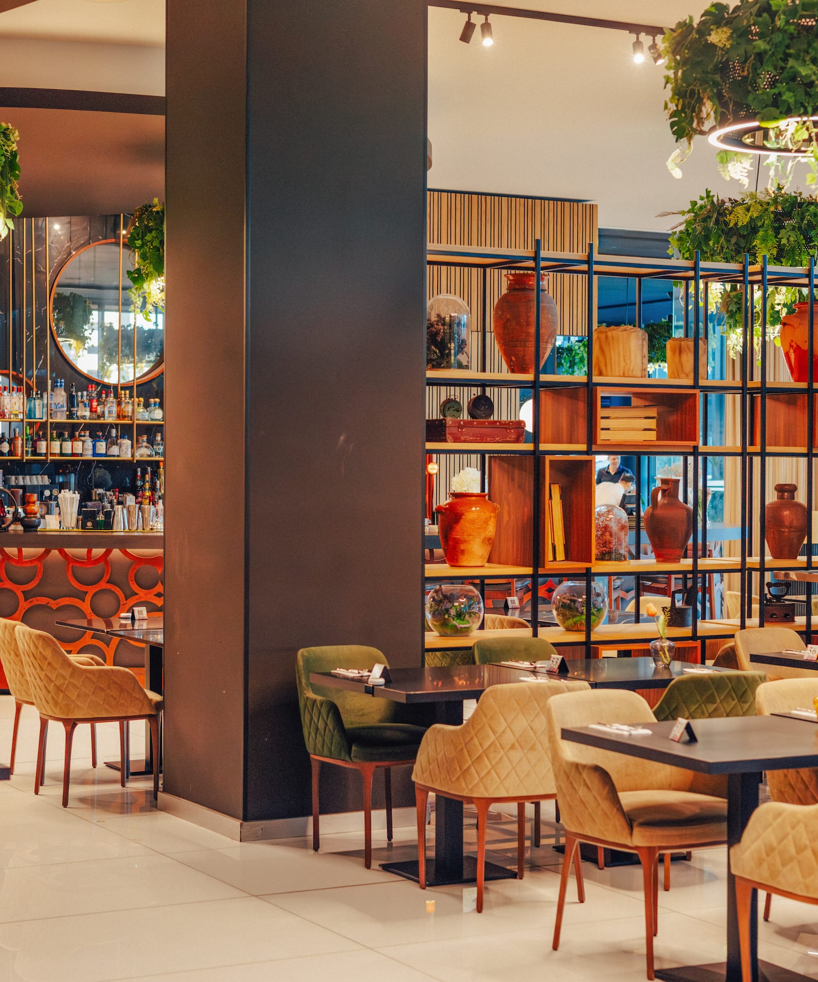 Stylish modern restaurant interior with dining tables, bar, decorative shelves, and hanging plants.