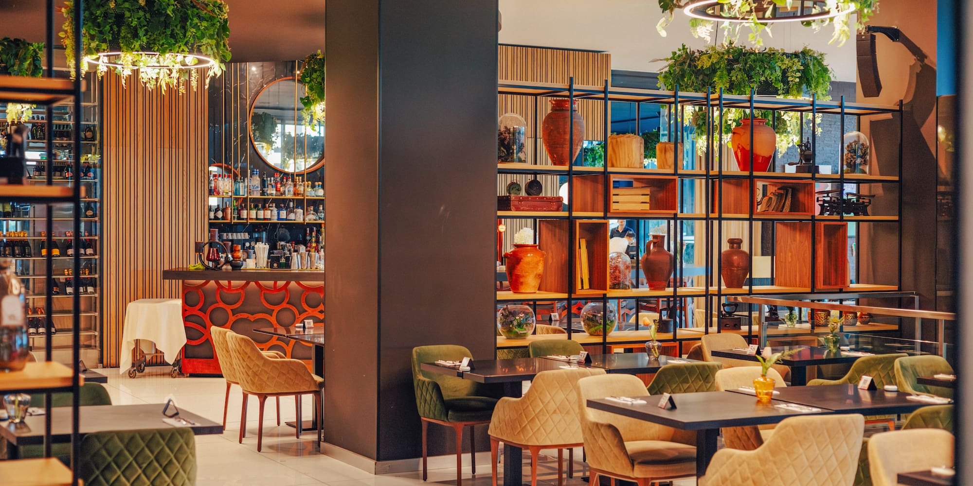 Stylish modern restaurant interior with dining tables, bar, decorative shelves, and hanging plants.