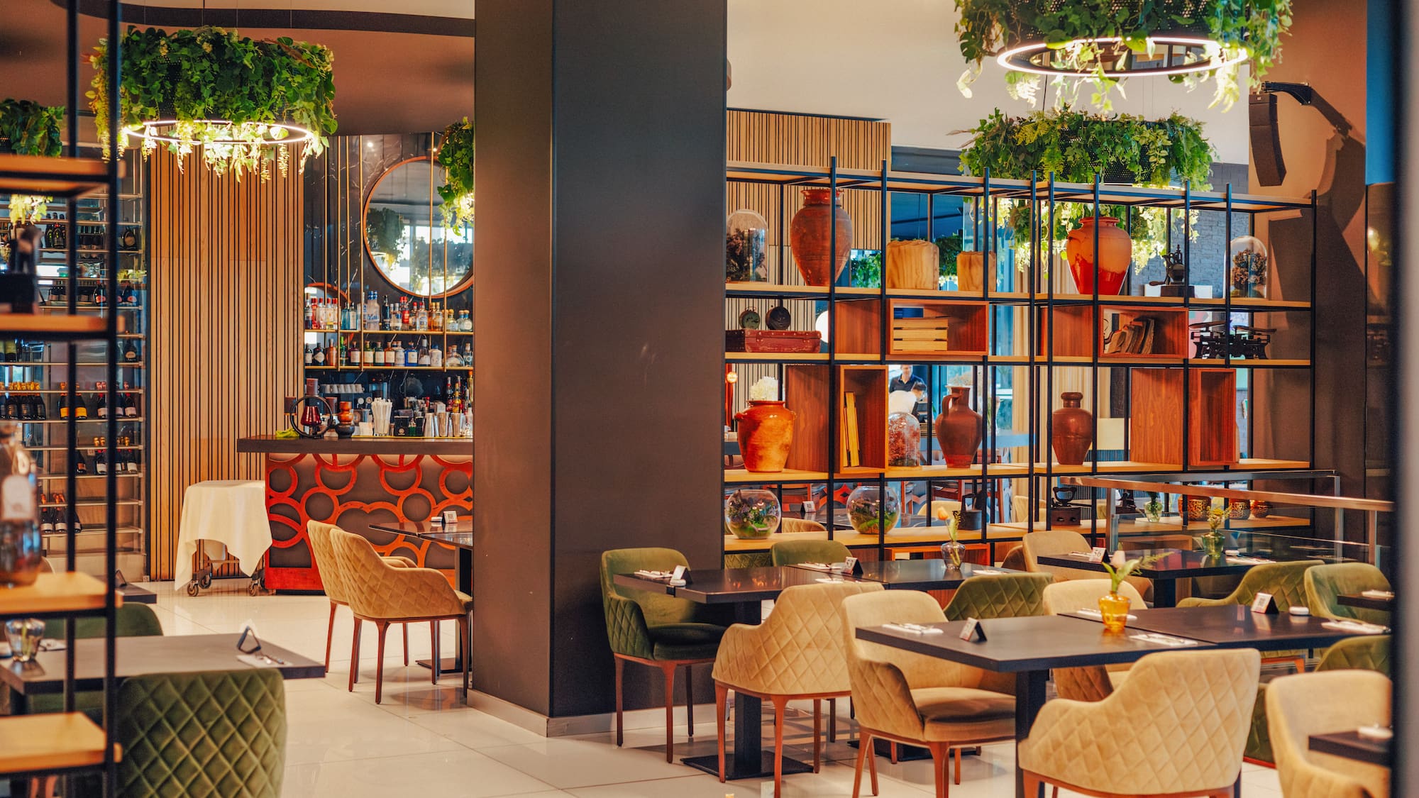 Modern restaurant dining room with bar, plush chairs, and hanging plants.