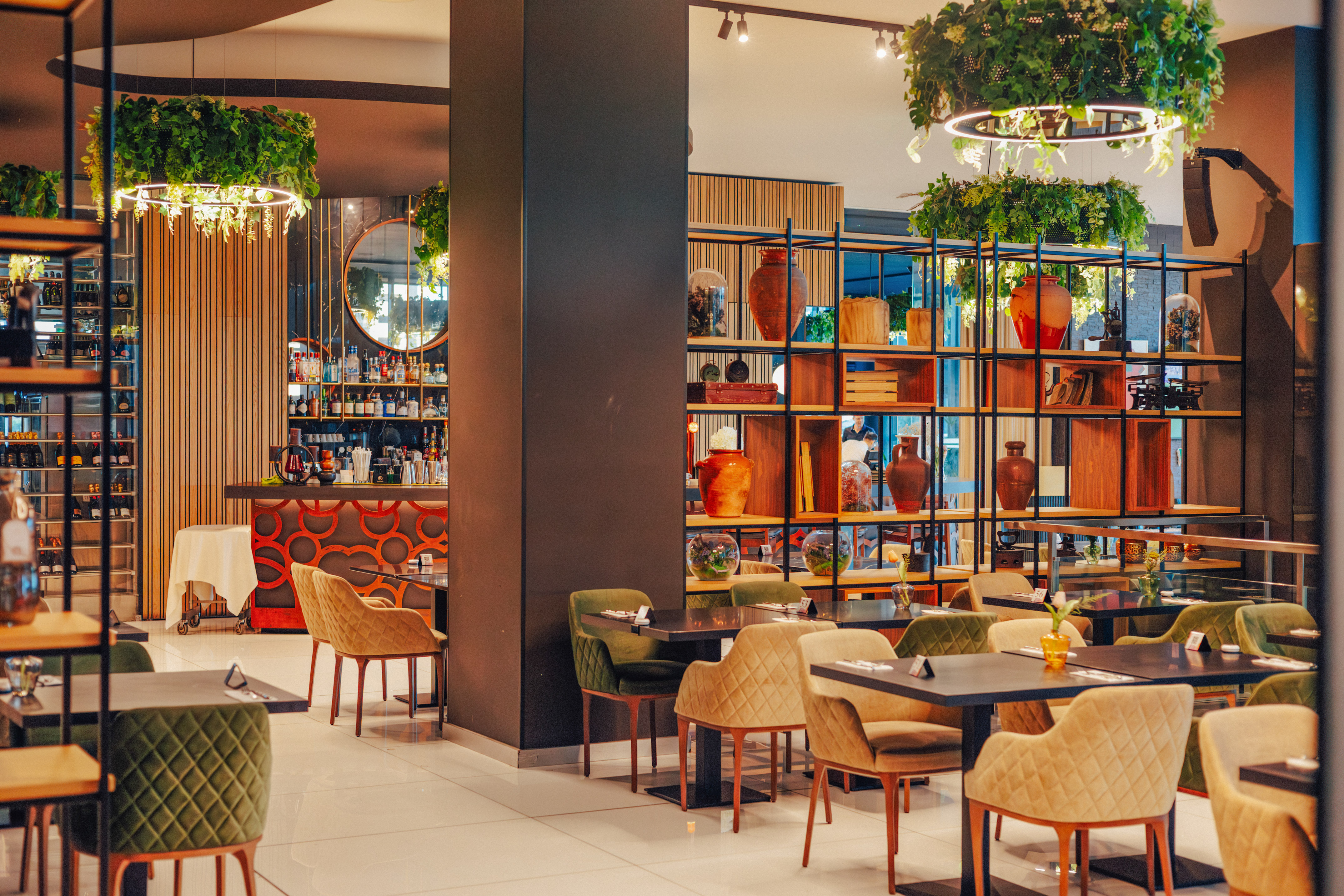 Modern restaurant dining room with bar, plush chairs, and hanging plants.