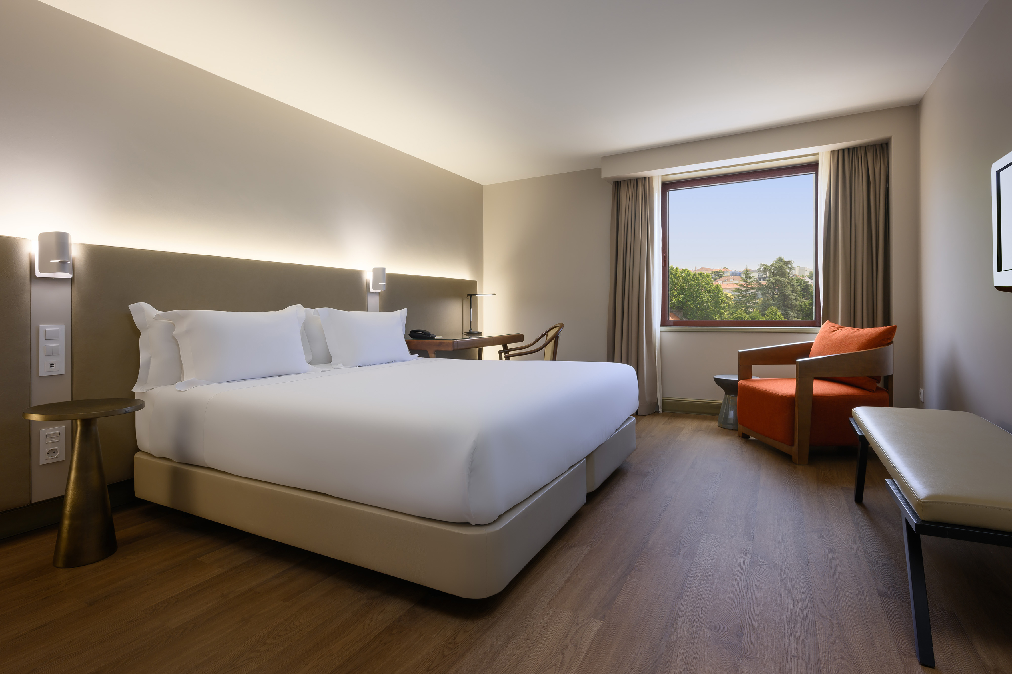 Modern hotel bedroom with king bed, desk, armchair, and window view.