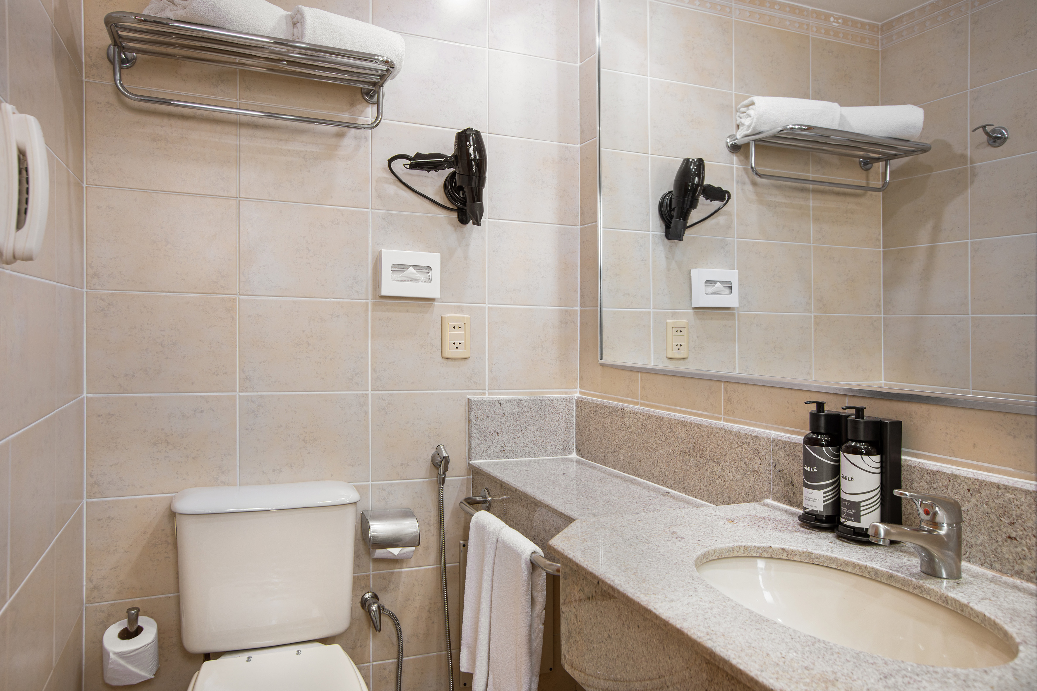 Beige tiled hotel bathroom, toilet, granite vanity, mirror, towels, hairdryer.