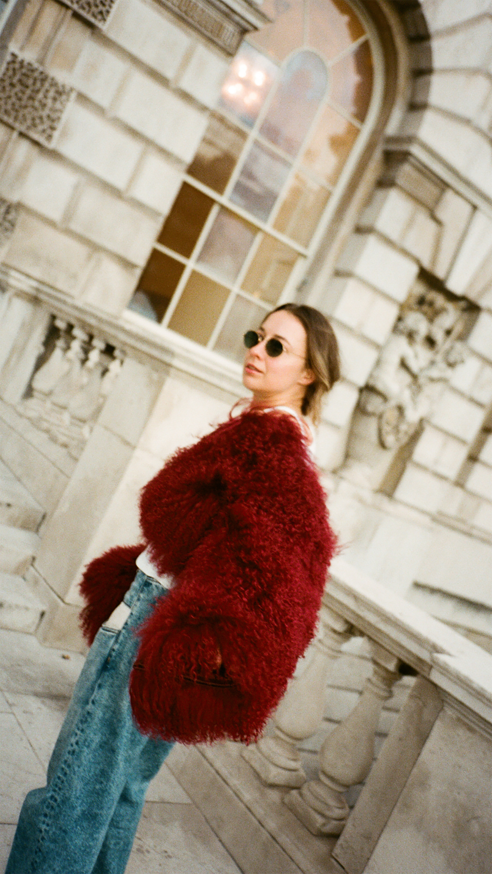 Woman in red fuzzy coat, blue jeans, sunglasses, against stone architecture.