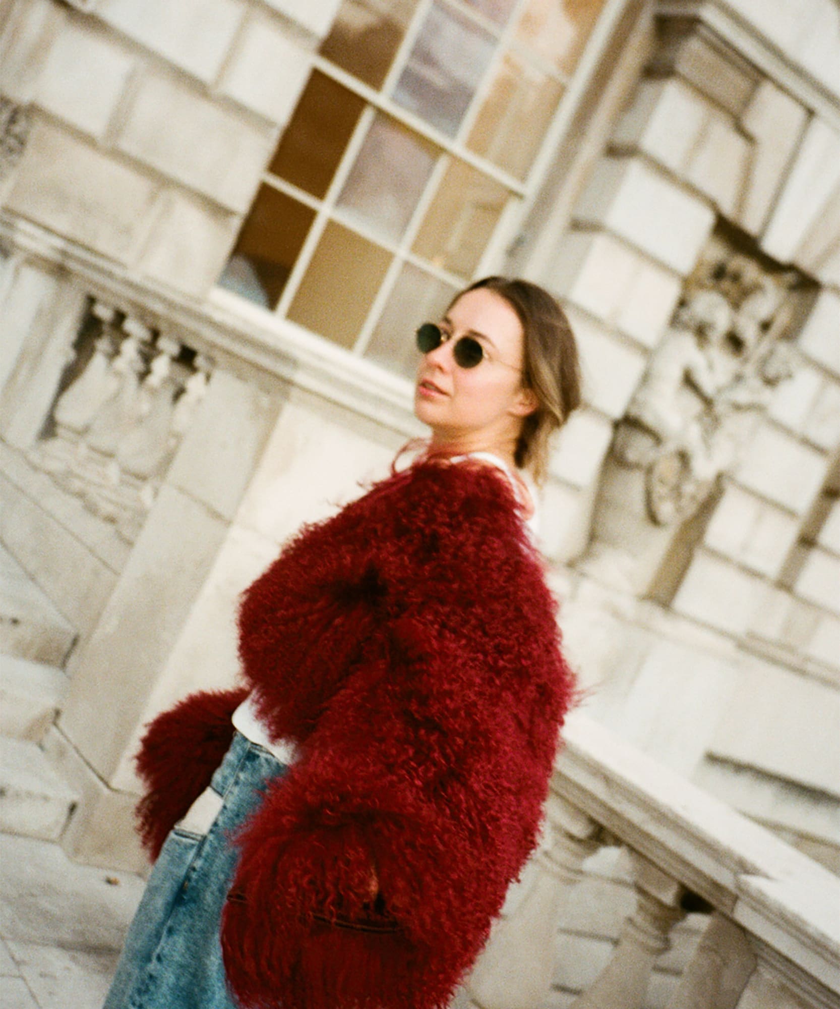 Woman in red fuzzy coat, blue jeans, sunglasses, against stone architecture.