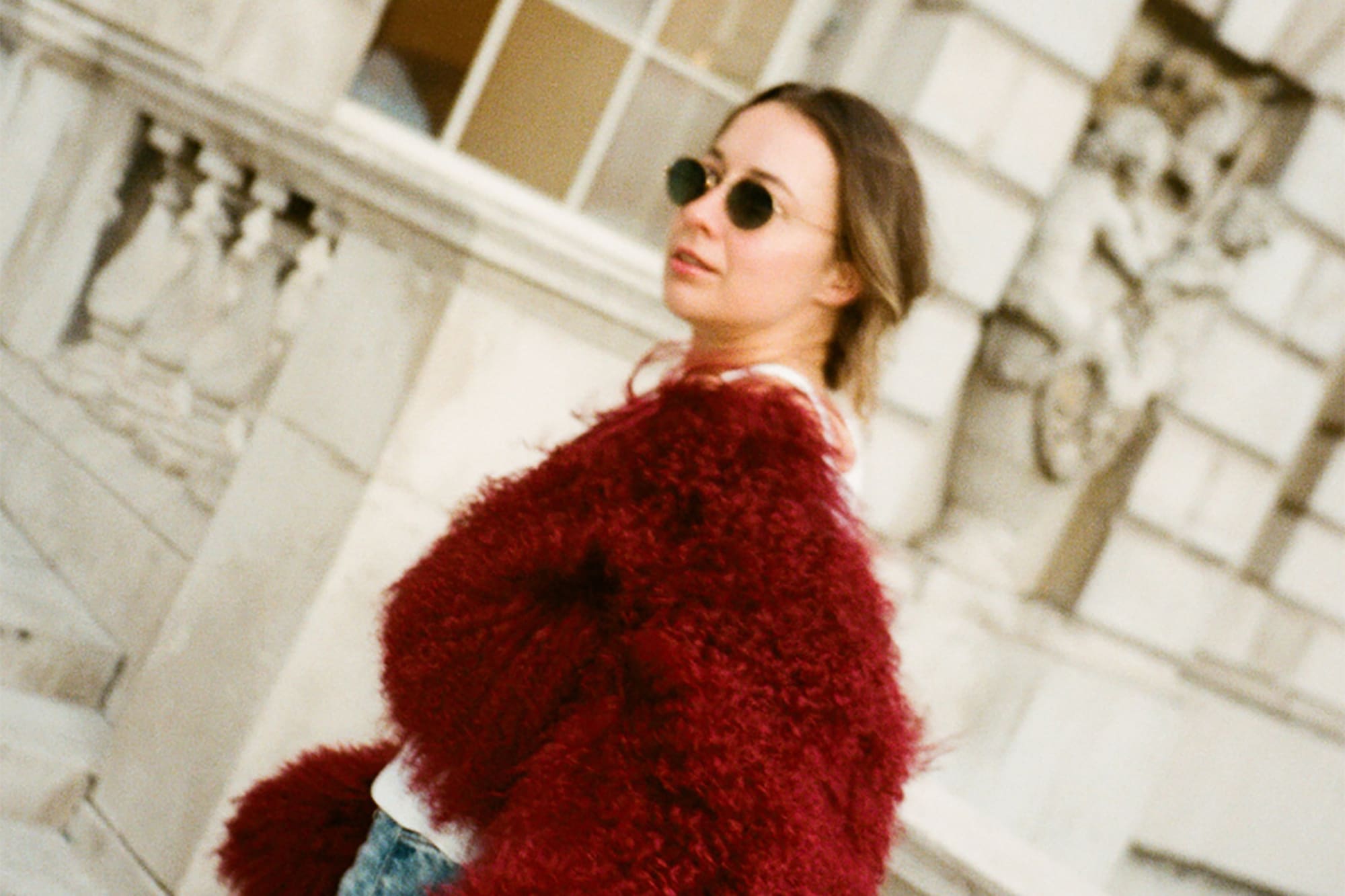 Woman in red fuzzy coat, blue jeans, sunglasses, against stone architecture.