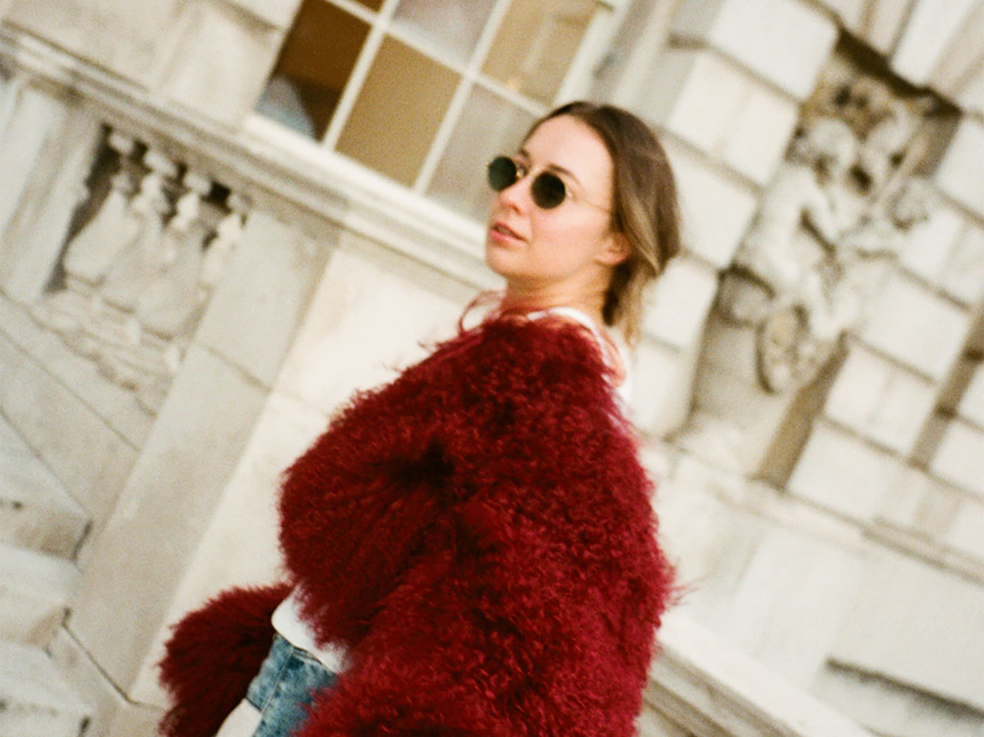 Woman in red fuzzy coat, blue jeans, sunglasses, against stone architecture.
