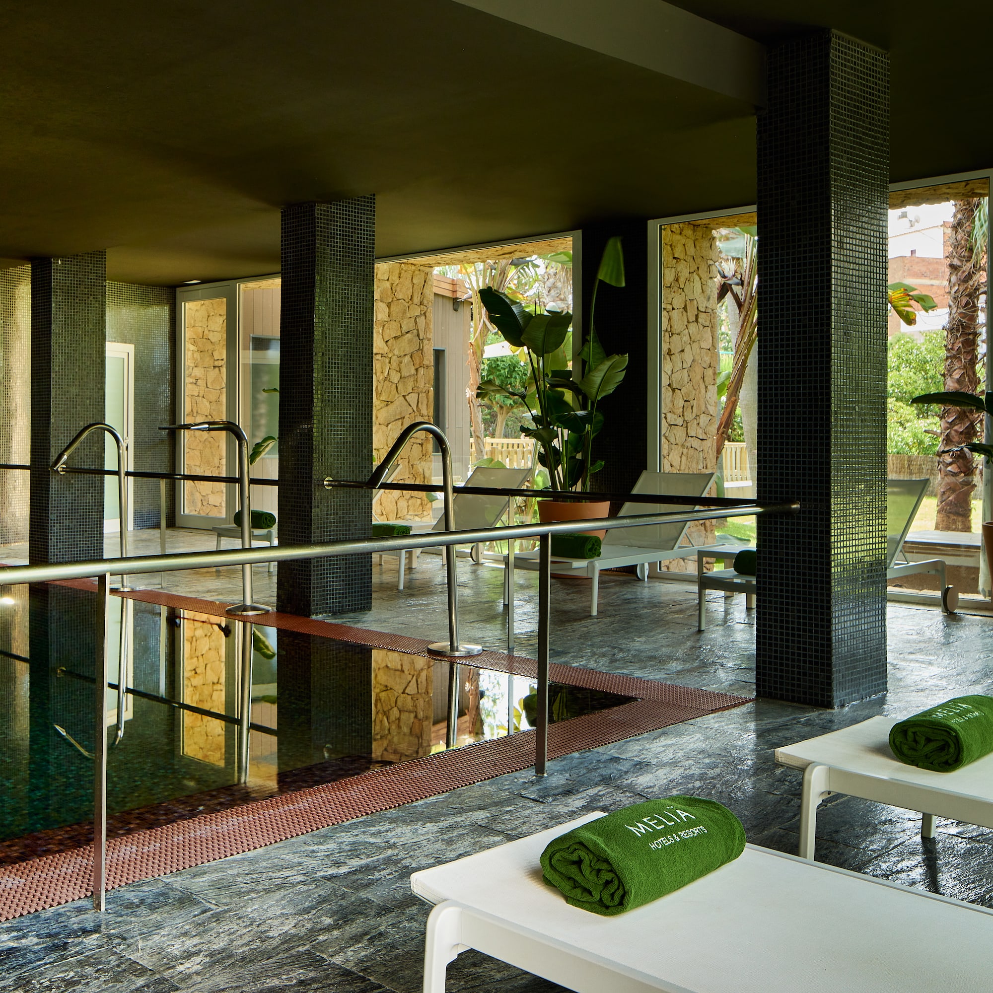 Piscina cubierta Meliá, tumbonas, toallas verdes y plantas tropicales.