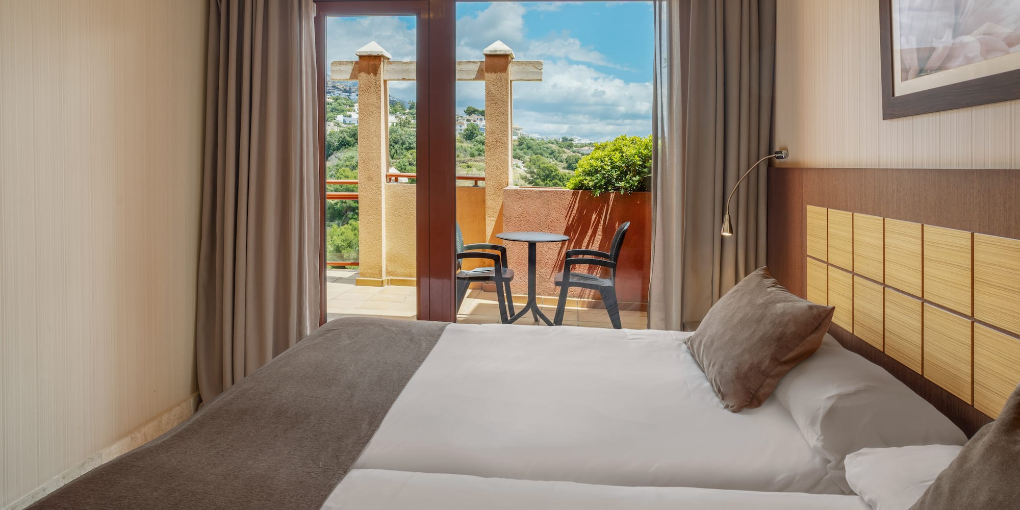 Hotel room with bed, private balcony, outdoor seating, and green hill view.