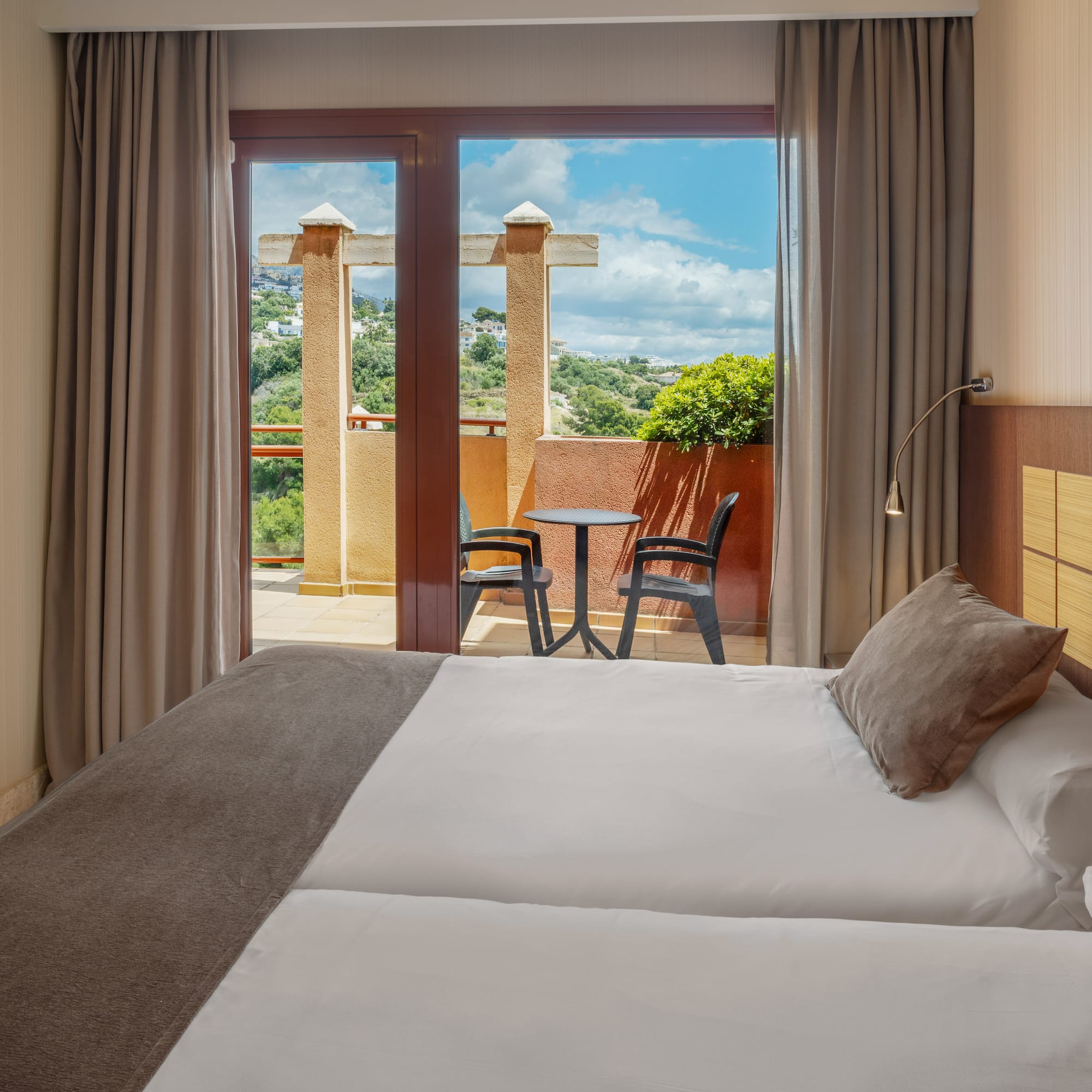 Hotel room with bed, private balcony, outdoor seating, and green hill view.