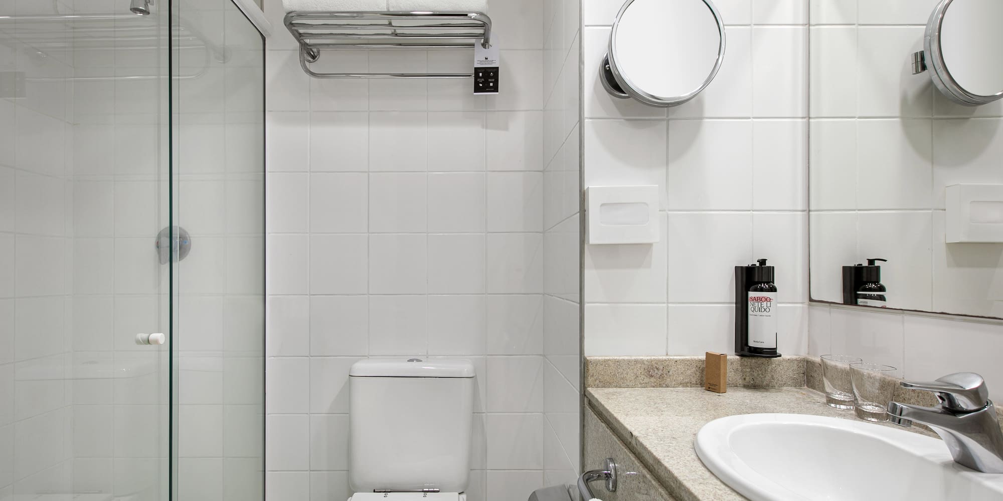 Clean hotel bathroom with white tiles, glass shower, toilet, and vanity.