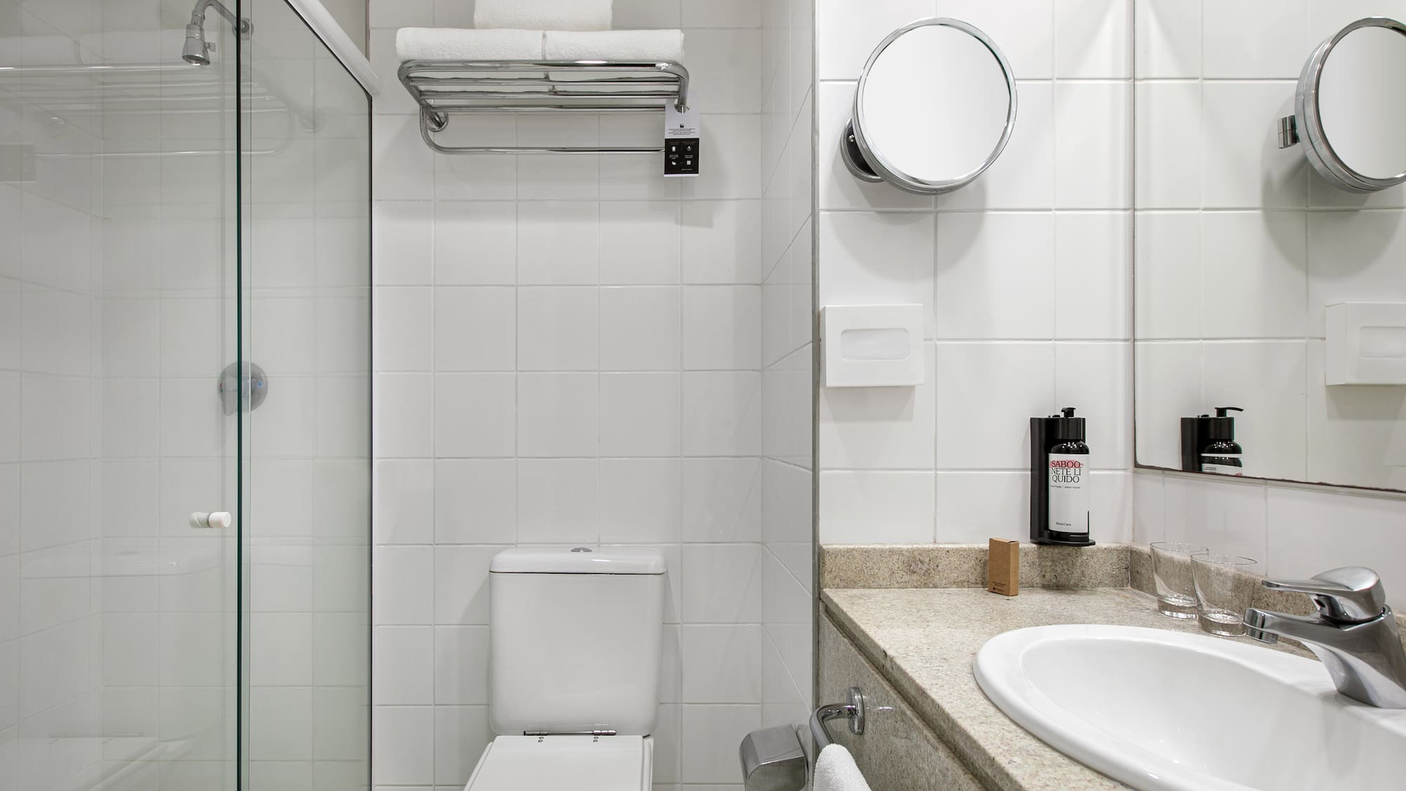 Clean hotel bathroom with white tiles, glass shower, toilet, and vanity.