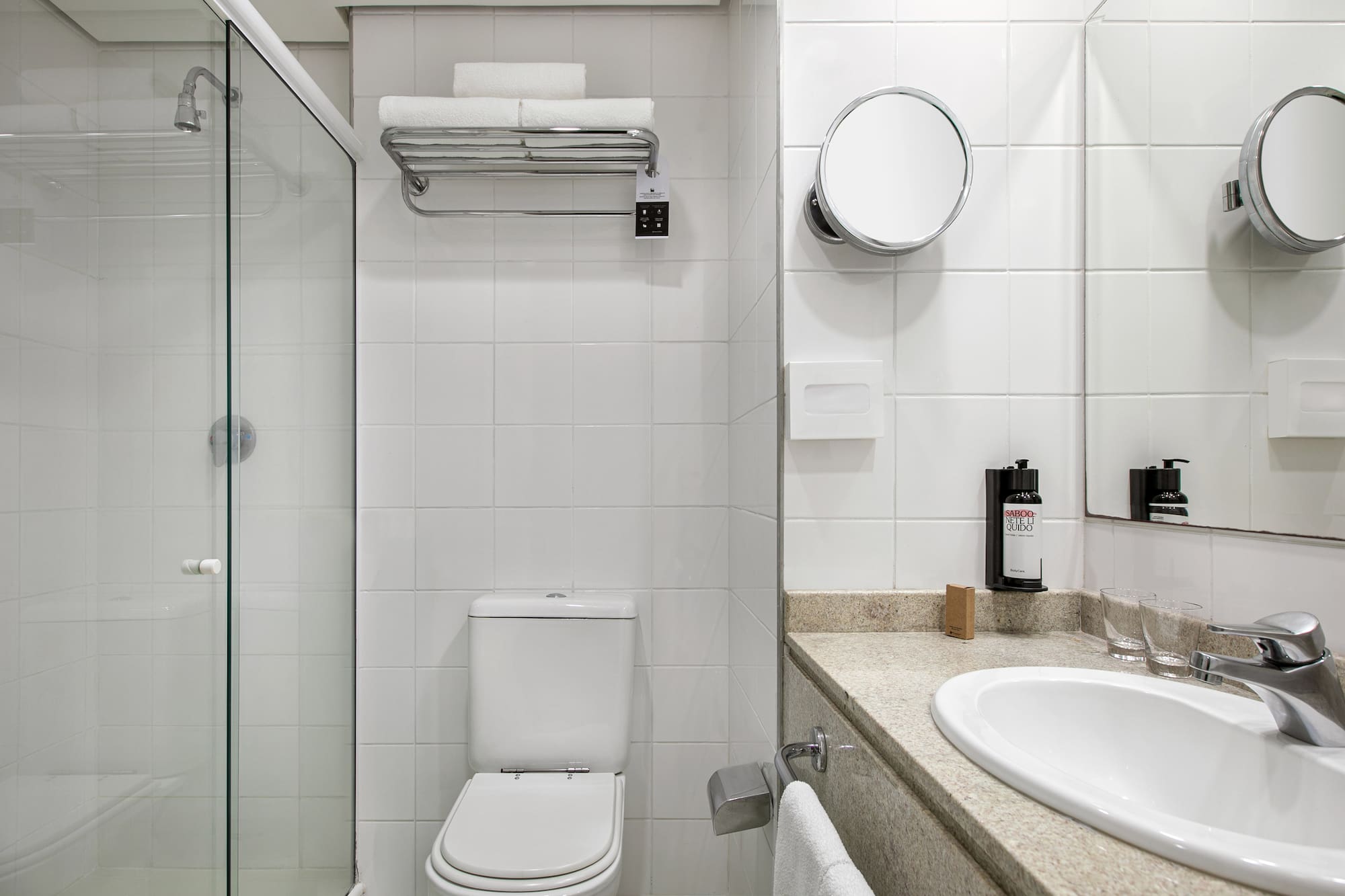 Clean hotel bathroom with white tiles, glass shower, toilet, and vanity.