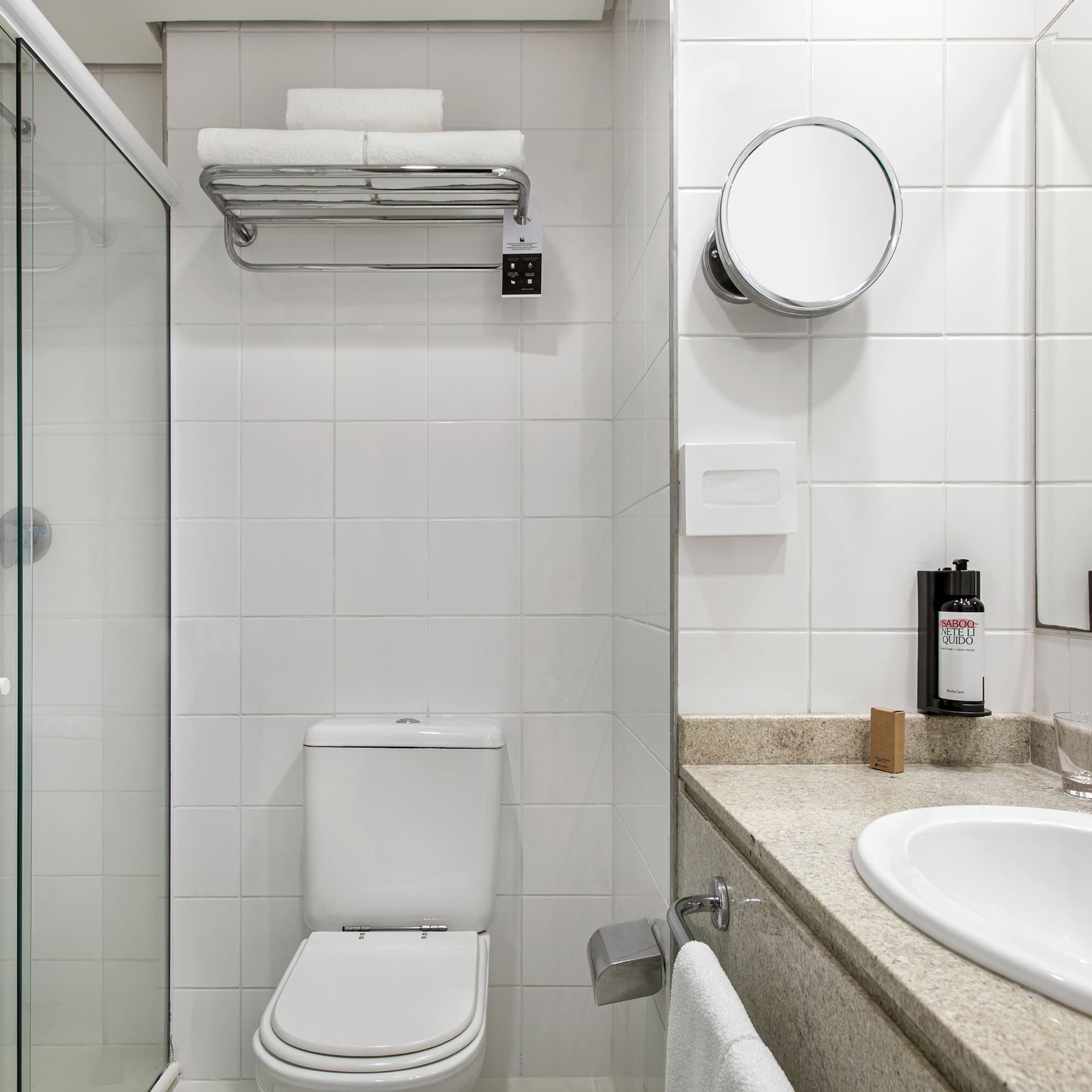 Clean hotel bathroom with white tiles, glass shower, toilet, and vanity.