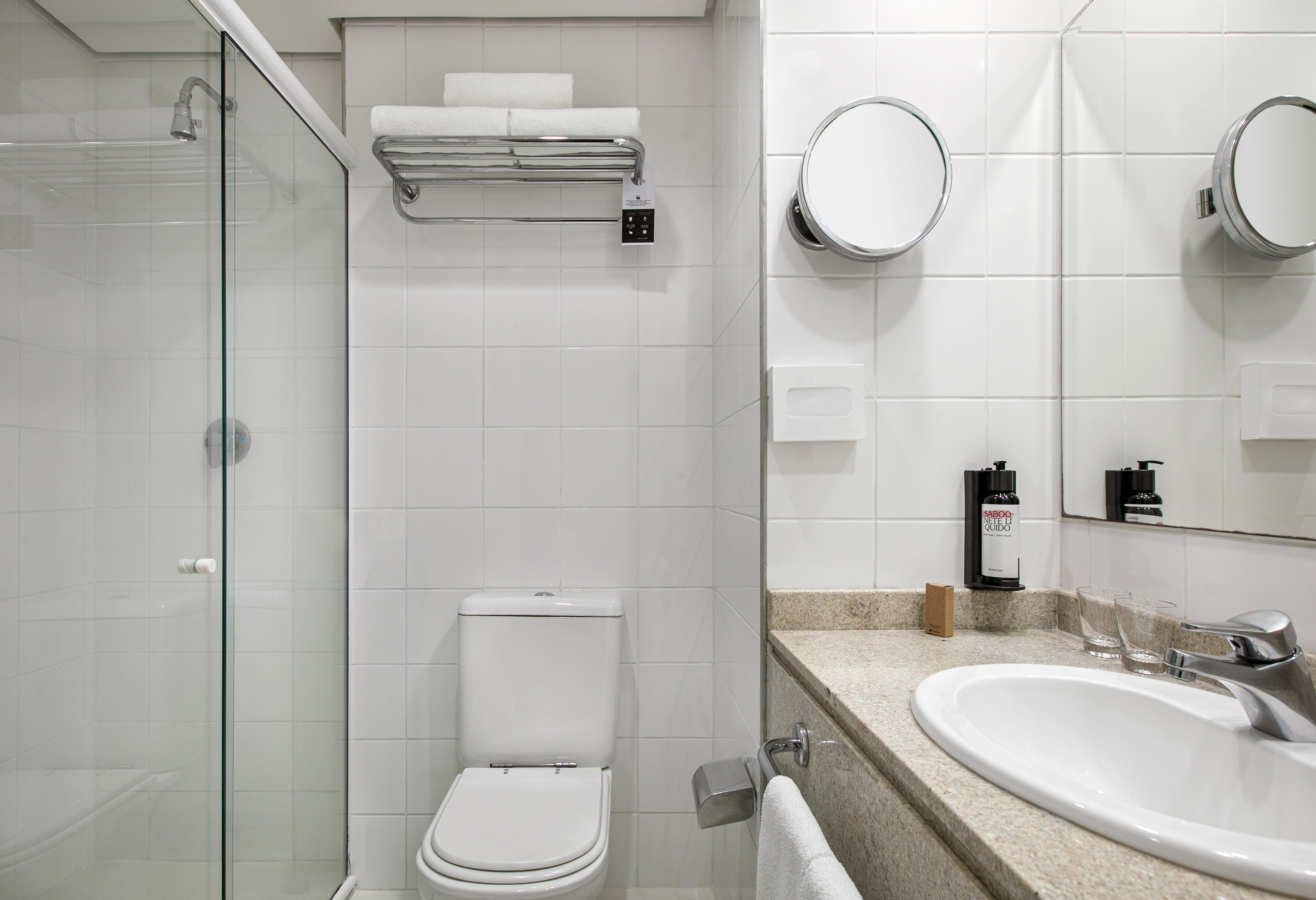 Clean hotel bathroom with white tiles, glass shower, toilet, and vanity.