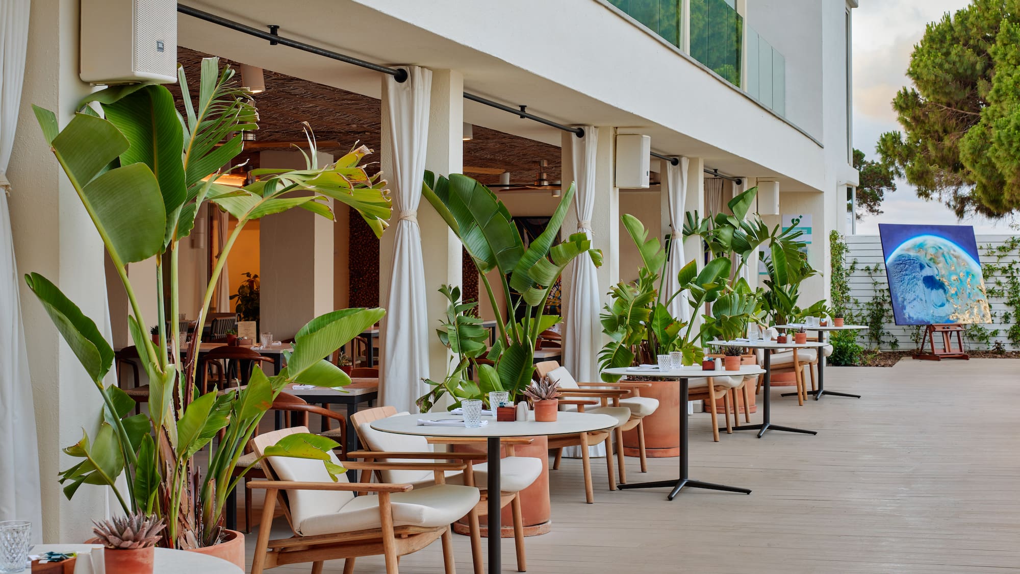 Modern outdoor dining area with tables, chairs, and lush tropical plants.