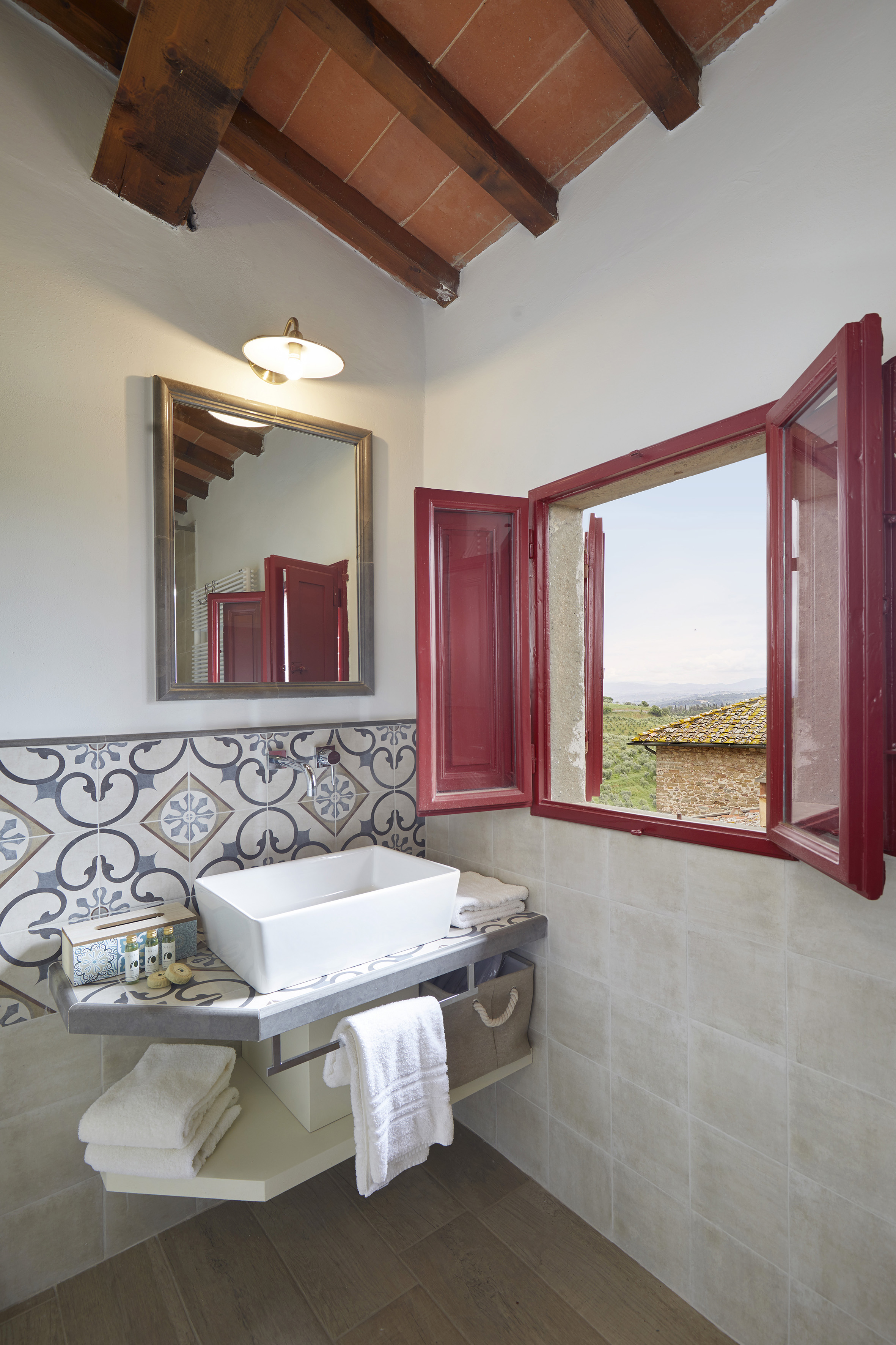 Tuscan bathroom: vessel sink, patterned tiles, wood beams, and red window with scenic view.